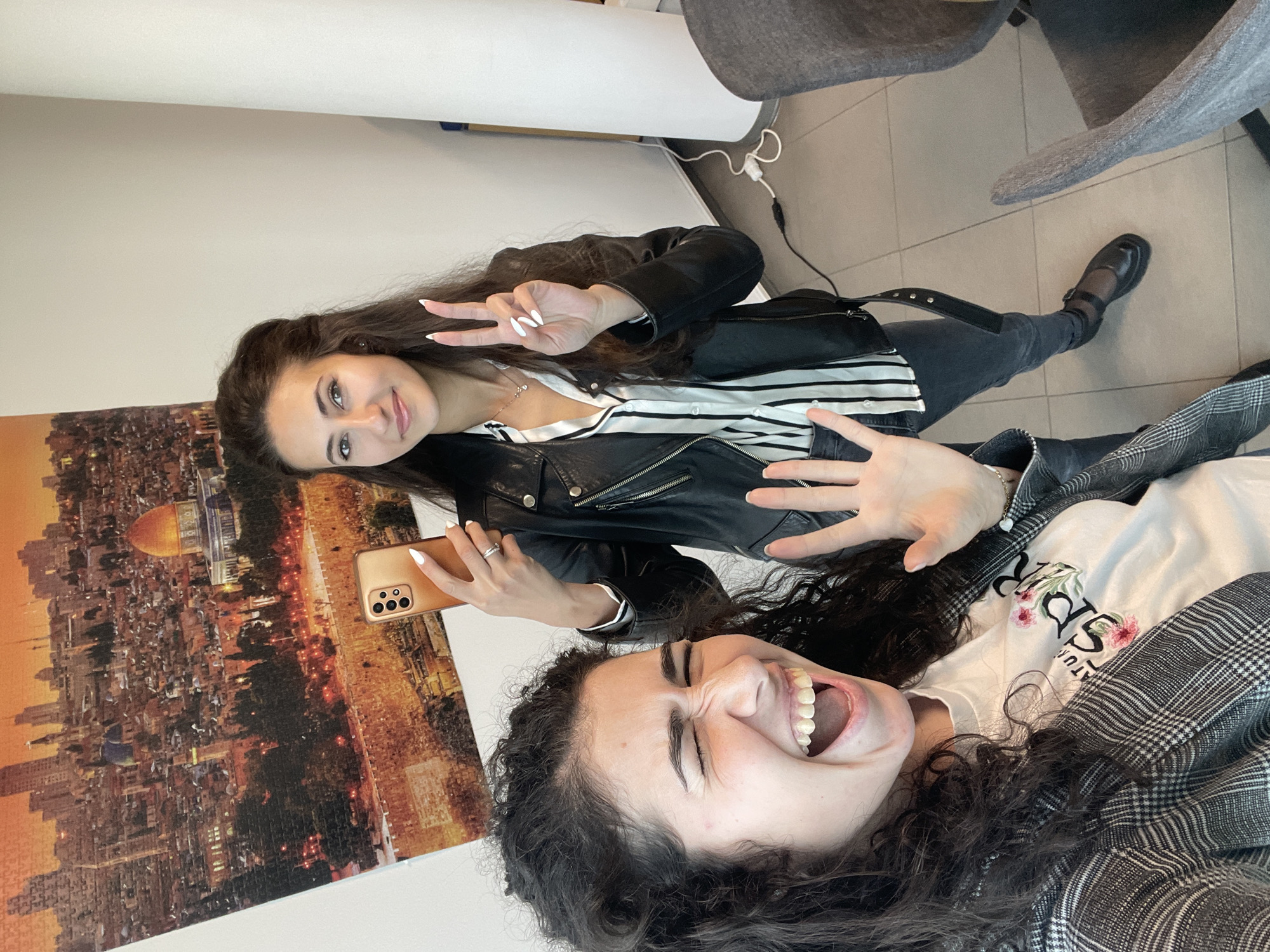 Two women are taking a selfie indoors. One woman, with dark curly hair, is lying down and laughing, wearing a white t-shirt with floral designs and a grey and white checkered jacket. The other woman, with long straight dark hair, is sitting up, holding a gold-colored phone and making a peace sign. She is wearing a black leather jacket over a black and white striped shirt, dark jeans, and black shoes. A black purse is strapped across her body. In the background, there is a puzzle featuring an aerial view of Jerusalem, a white wall, a white cylindrical object, and grey chairs. It is not possible to determine if the location is in Sofia, Bulgaria based on the visual information provided.