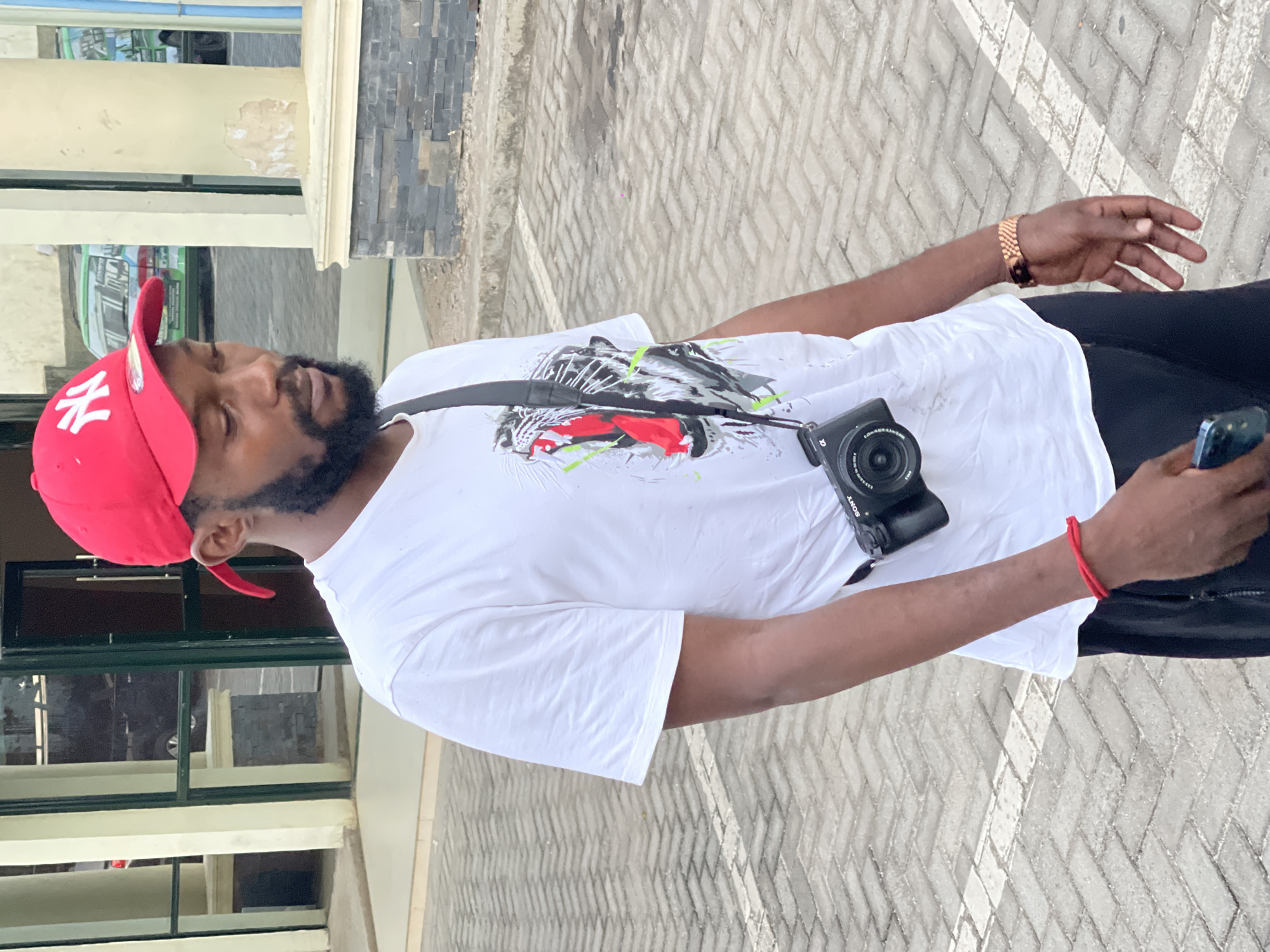 The image, rotated 90 degrees clockwise, captures a casually dressed man standing outdoors in what appears to be Abuja, Nigeria. The man, with dark skin and a neatly trimmed beard, is seen from the waist up, facing slightly to his right. He wears a bright red baseball cap with white "NY" lettering on the front, and a plain white t-shirt featuring a stylized graphic of a snarling feline or tiger head in black, white, red, and neon green.
A black camera, clearly identifiable as a "SONY" brand with lens details like "0.2M/0.65ft" visible, hangs across his body on a black strap. On his left wrist, he wears a gold-colored link wristwatch, and on his right wrist, a simple red string bracelet. He holds a dark-colored smartphone in his right hand.
The background consists of an outdoor setting with light grey interlocking paving blocks arranged in a herringbone pattern. Behind him, parts of a building are visible, including a cream-colored column or pillar, dark grey stacked stone cladding, and windows with dark green frames. The lighting is bright, suggesting daytime. The man's expression is neutral, and his gaze is directed downwards, possibly at his phone or the ground. The overall mood is casual and observational.