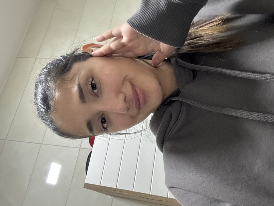 A young woman in a grey hoodie looks directly at the camera with a slight smile. Her hand is near her ear, possibly adjusting her hair. Her dark hair is pulled back, revealing her forehead. The background includes a tiled wall and the edge of a white desk or cabinet. The overall lighting is bright and even, giving the image a clean, casual feel.