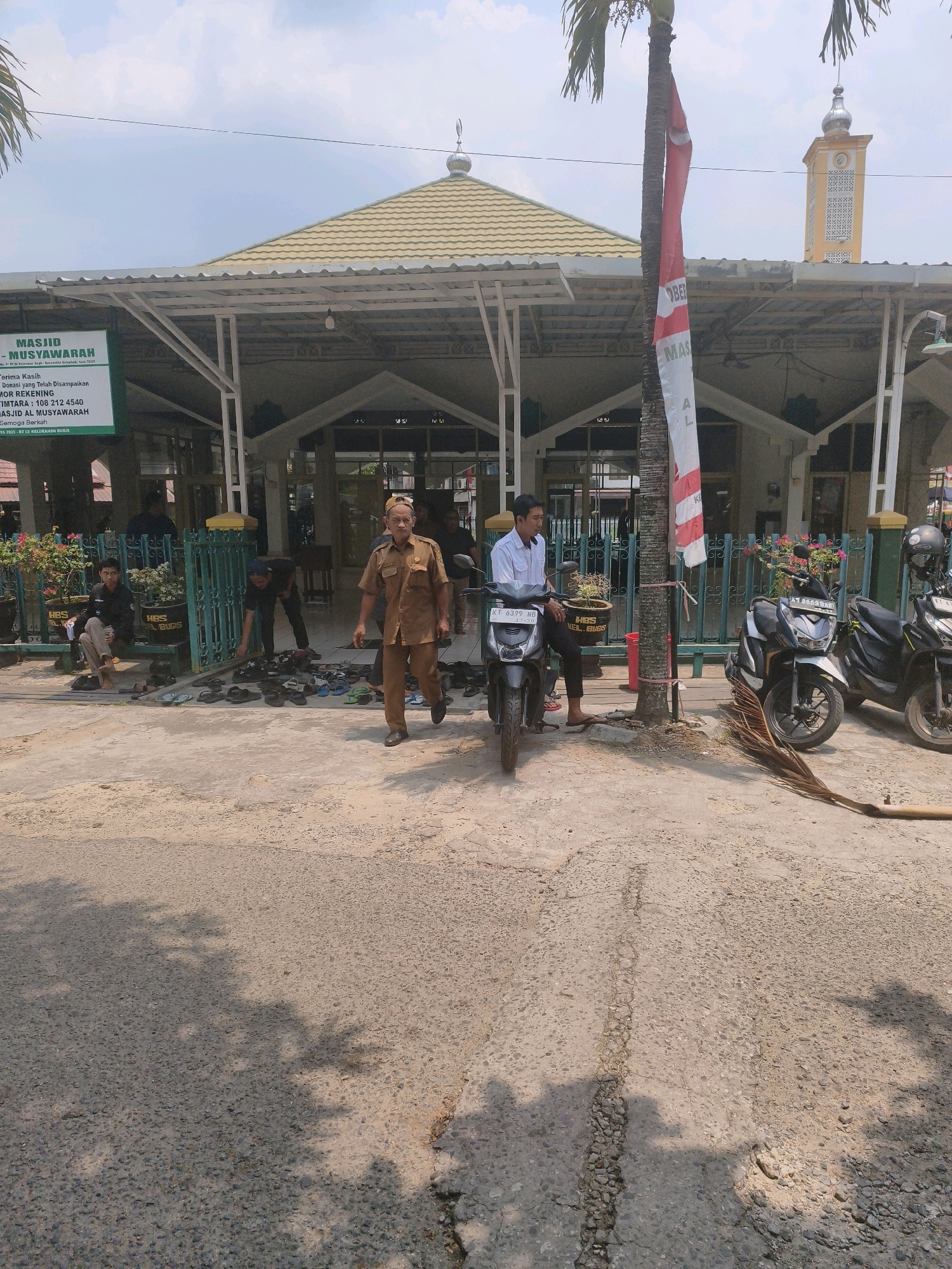 This is a medium shot, eye-level, outdoor, daylight photograph of a mosque named Masjid Al Musyawaroh in the city of Samarinda, Indonesia. The mosque has a yellow tiled roof with white trim. There is a tall, narrow minaret to the right of the mosque, also yellow with white trim. In front of the mosque is a covered walkway supported by white columns. A red and white flag is tied to a palm tree in front of the mosque. Several people are visible. One man in a tan uniform walks towards the camera on the right side of the frame. Another man in a white shirt and black pants sits on a motorcycle to the left of the frame. Several other people are visible in the background, some sitting and some standing. There are many pairs of sandals and shoes on the ground in front of the mosque, suggesting people have removed them before entering. There are also two motorcycles parked in front of the mosque, and a broom made of palm fronds lies on the ground. The ground in the foreground is a rough, gray asphalt surface with some cracks and pebbles. Shadows of trees are cast on the ground. A green fence is visible along the front of the mosque's grounds. A sign in front of the mosque reads, "- MASJID AL MUSYAWAROH - TERIMA KASIH - MOR REKENING TIMTARA: 108 212 4540 - MASJID AL MUSYAWARAH - Semoga Barkah". The sign indicates the mosque is located in Kelurahan Bugis, RT 03.