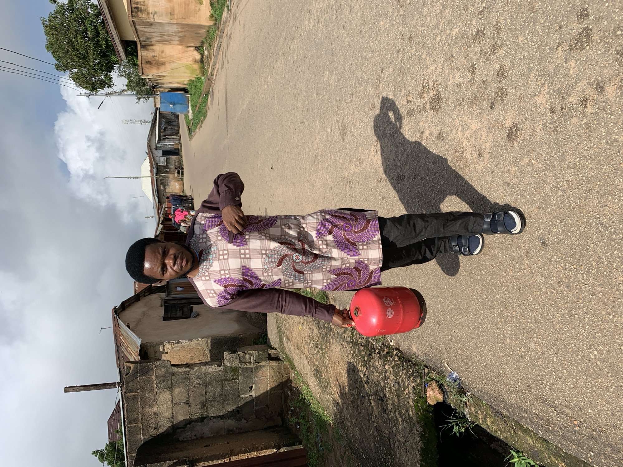 A young Black male walks towards the camera down a paved road in Jos, Nigeria. He is wearing a brown long-sleeved shirt with a purple and white patterned design, black pants, and dark sandals. He is carrying a red propane gas cylinder in his left hand. The road is lined with simple, aging buildings with corrugated metal roofs on the left and a more modern building with a tan wall on the right. There are power lines and a cell tower visible in the background. The sky is partly cloudy.