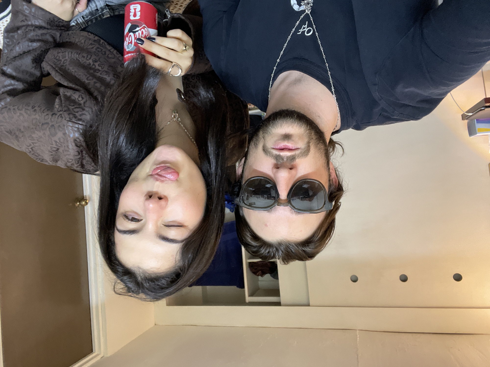 The image shows a selfie of two individuals. A woman is holding a can of Coca-Cola. She is winking and sticking out her tongue. She is wearing a brown patterned jacket, black nail polish, and jewelry. Next to her is a man with sunglasses, a beard, and a black shirt with a silver chain. The location appears to be indoors with a door and a cabinet in the background. There is no information about this image being in Naucalpan de Juárez, Mexico.