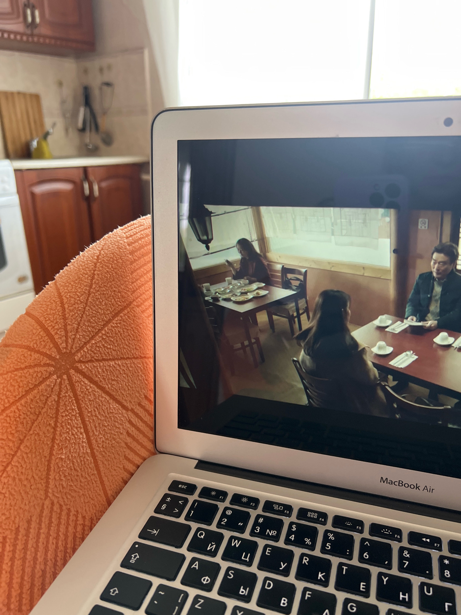 An overhead close-up shot shows a MacBook Air laptop open and displaying a scene from a television show or movie. In the foreground, the keyboard of the laptop is visible, as is an orange textured blanket or cushion. The laptop screen shows three people seated at tables in what appears to be a restaurant. One woman is seated at a table on the left, looking down at her phone. Two other people, a man and a woman, are seated at a table on the right, facing each other. The man is looking at the woman and holding a small object in his hands. The background of the room appears to be paneled in wood, and a window shows an outdoor scene. In the far background of the overall image, a kitchen area with wooden cabinets and a countertop is visible. The scene is indoors and appears to be in a domestic setting, possibly a home or a small eatery, and is not identifiable as being located in the city of Morozovsk, Russia.