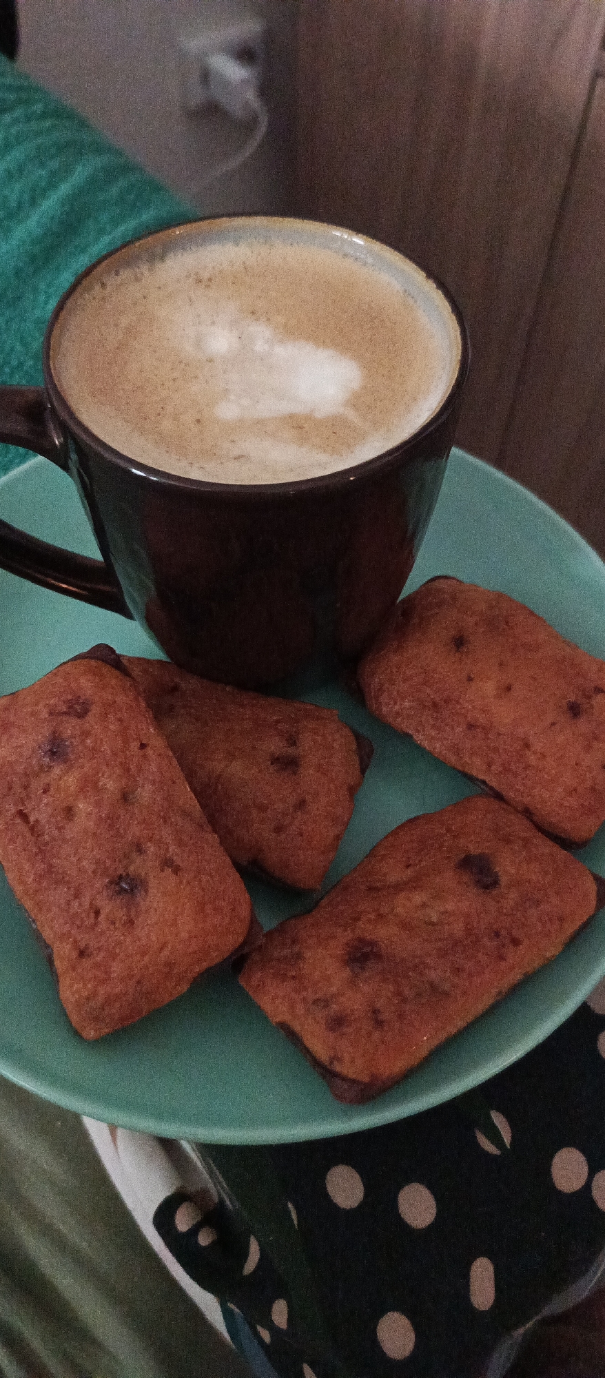 A dark brown mug of coffee with frothy milk sits on a teal plate, next to a pile of four rectangular baked goods. The coffee has a light brown frothy surface with a distinct white swirl. The plate is round and has a slight lip. The baked goods appear to be some type of small cakes or cookies, with a reddish-brown color and visible darker speckles, likely chocolate chips or dark fruit. One of the baked goods is partially obscured by the mug, and the other three are arranged in front of it on the plate. The plate itself is set on a surface that appears to be a dark fabric with a white polka dot pattern. To the left of the plate, a textured teal fabric, possibly a blanket or towel, is visible. The background is blurred but suggests an indoor setting with a wall and a light fixture. The overall scene depicts a snack or breakfast setting. No specific location in Bugry, Russia can be determined from the image.