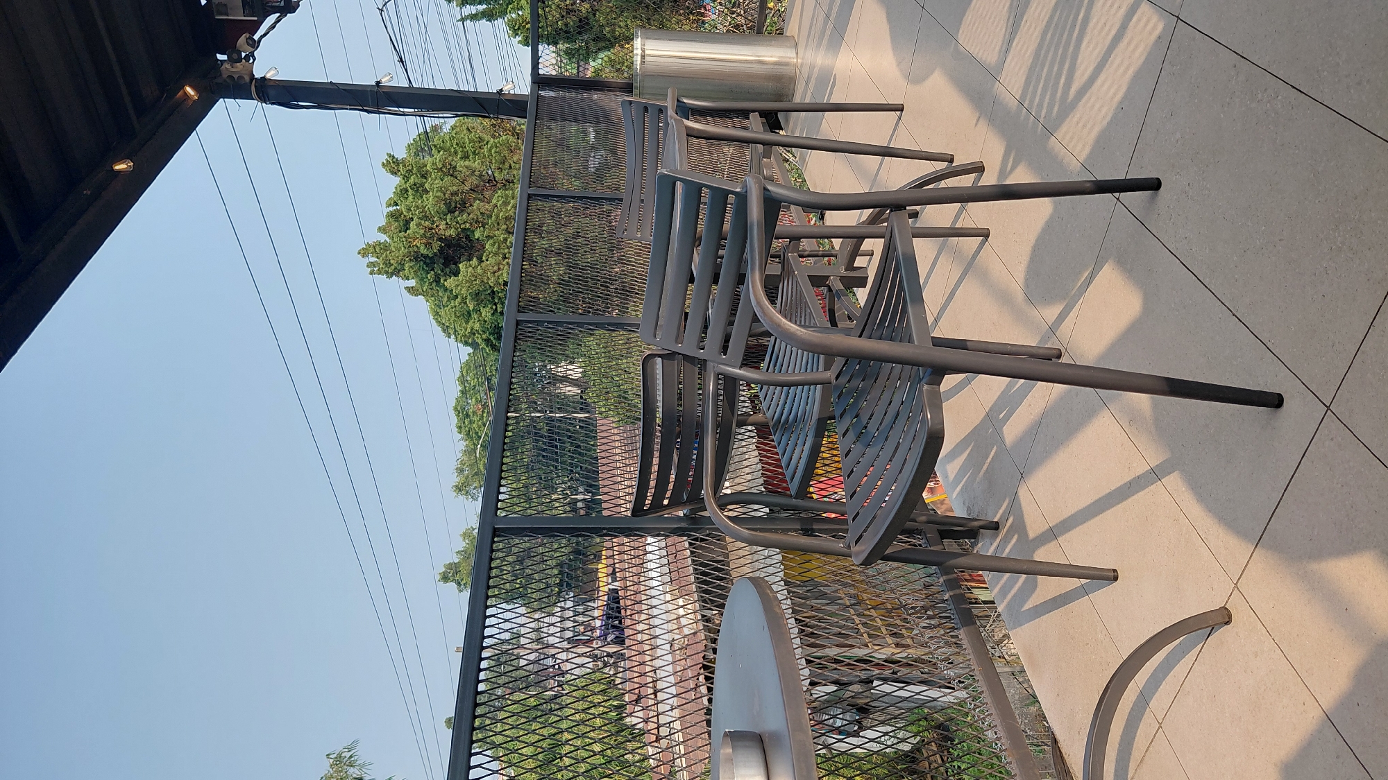The image shows an outdoor seating area with several dark gray metal chairs and a round table. The furniture is placed on a tiled patio, with a black metal fence in the background. Beyond the fence, there are rooftops, trees, and power lines, suggesting an urban or semi-urban setting. The sky is clear and blue, and the sunlight casts long shadows on the patio, indicating it is likely late afternoon or early morning. The location is identified as Karanglewas, Indonesia. There are no people visible in the image, and the scene is static.
