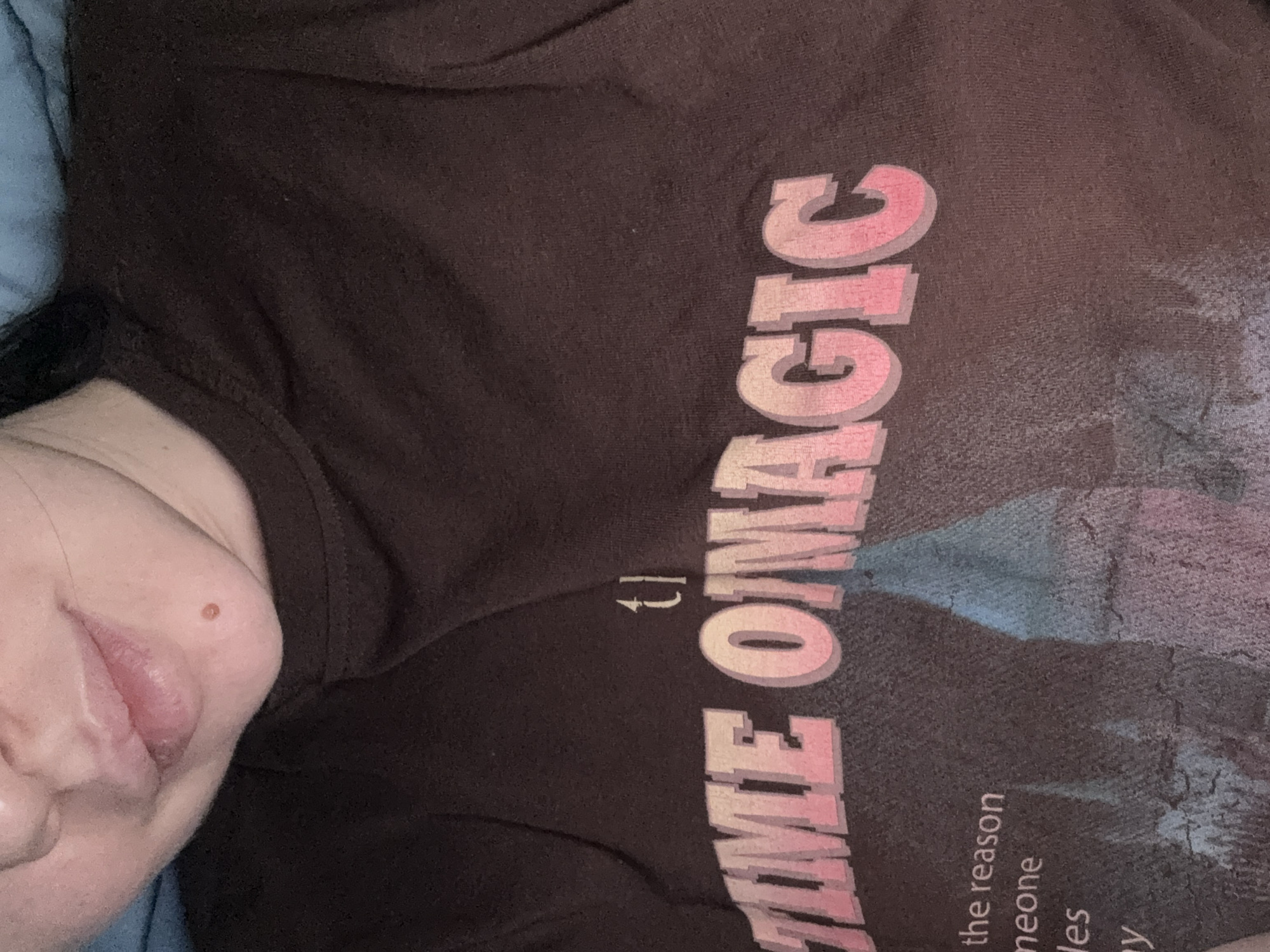 The image shows a person wearing a brown t-shirt with the words "TIME OMAGIC" printed on it in pink. The person's face, specifically the mouth and chin area, is visible in the lower left corner of the frame. Part of a light blue or grey surface, possibly a pillow or blanket, is visible in the top left corner. There is no location information, particularly related to Mytishchi, Russia, present or derivable from the image.