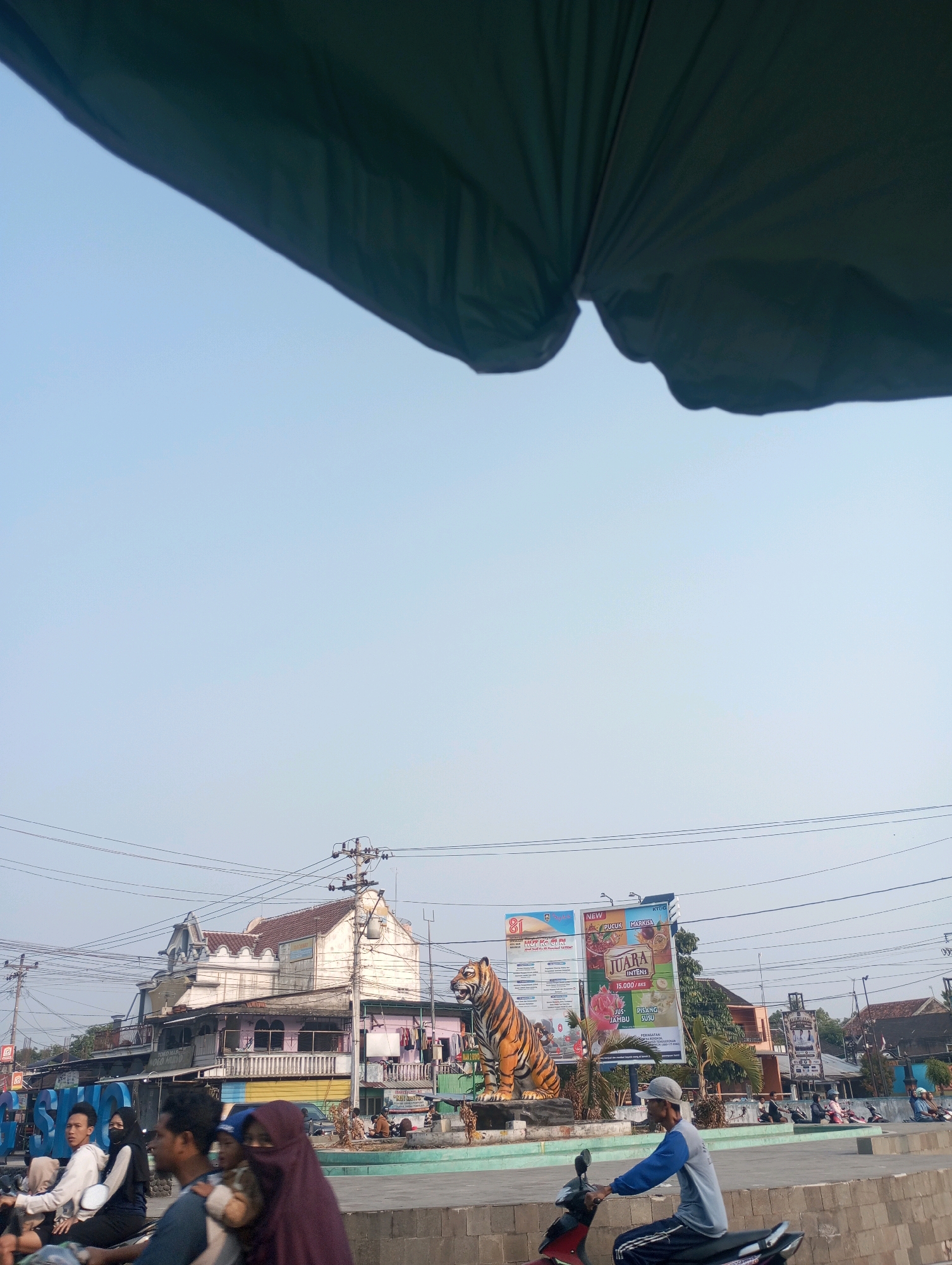 In the city of Simo, Indonesia, a street scene unfolds under a bright, clear sky. Dominating the central view is a large, lifelike statue of a tiger, its orange and black stripes sharply defined. To the left of the tiger is a large, blue, three-dimensional sign spelling out "GOSONG". Behind the tiger and sign is a multi-story building with a faded pink facade and an ornate white roofline. Several billboards are visible, including one advertising "JUARA INTENS" juice with images of dragon fruit and bananas. The foreground shows several people riding motorcycles, including a man in a blue and gray long-sleeved shirt and a cap, and a woman wearing a dark purple headscarf. A dark green tarp or awning is visible in the upper portion of the image, casting a shadow. The scene appears to be a public square or intersection, with power lines crisscrossing the sky and other buildings and people visible in the background.