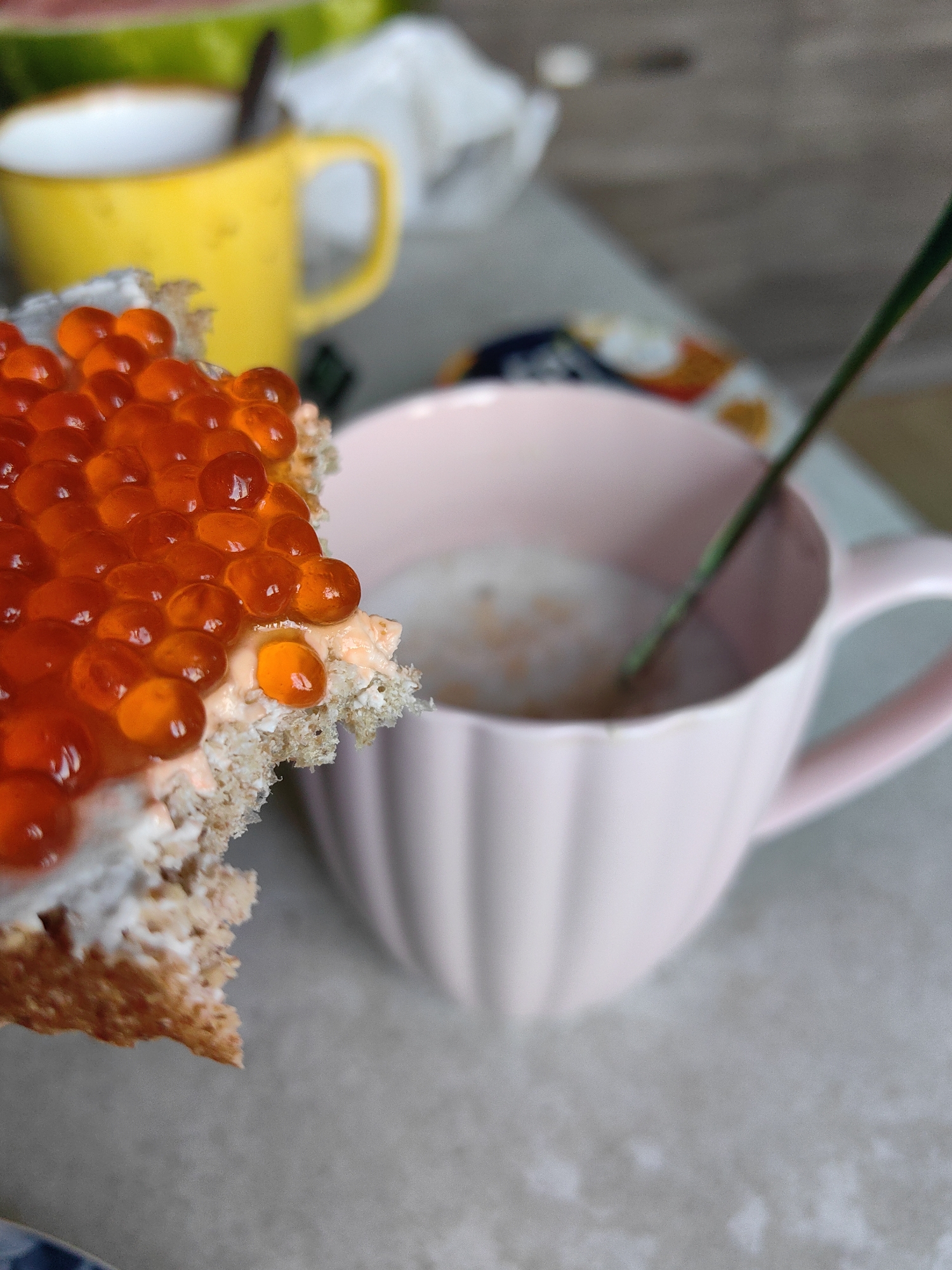A close-up, overhead shot shows a piece of bread with red caviar and cream cheese, held in the foreground. Behind it, a pink ceramic mug filled with a light-colored liquid and a green spoon is slightly out of focus. Further in the background, a yellow mug and a blurred slice of watermelon are visible. The setting appears to be a table, possibly at a residential location in Fanipal, Belarus.