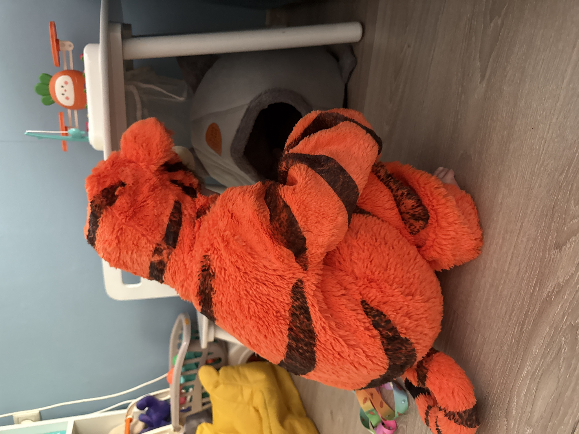A stuffed tiger toy is in the foreground, facing away from the camera and to the right. It is orange with black stripes and has its back and tail visible. The tiger is sitting on a wooden floor. In the background, there is a light-colored pet bed or den with a dark opening. Behind that, a white table with a small toy on it, which appears to be a scale with a carrot-shaped figure. To the left of the tiger, there is a yellow object, possibly a blanket or another stuffed toy, and a toy basket filled with colorful items. The wall in the background is light blue. The scene appears to be indoors, likely in a child's room or a play area. There are no people or animals visible in the image, apart from the stuffed tiger. The location is not identifiable as Moscow, Russia.