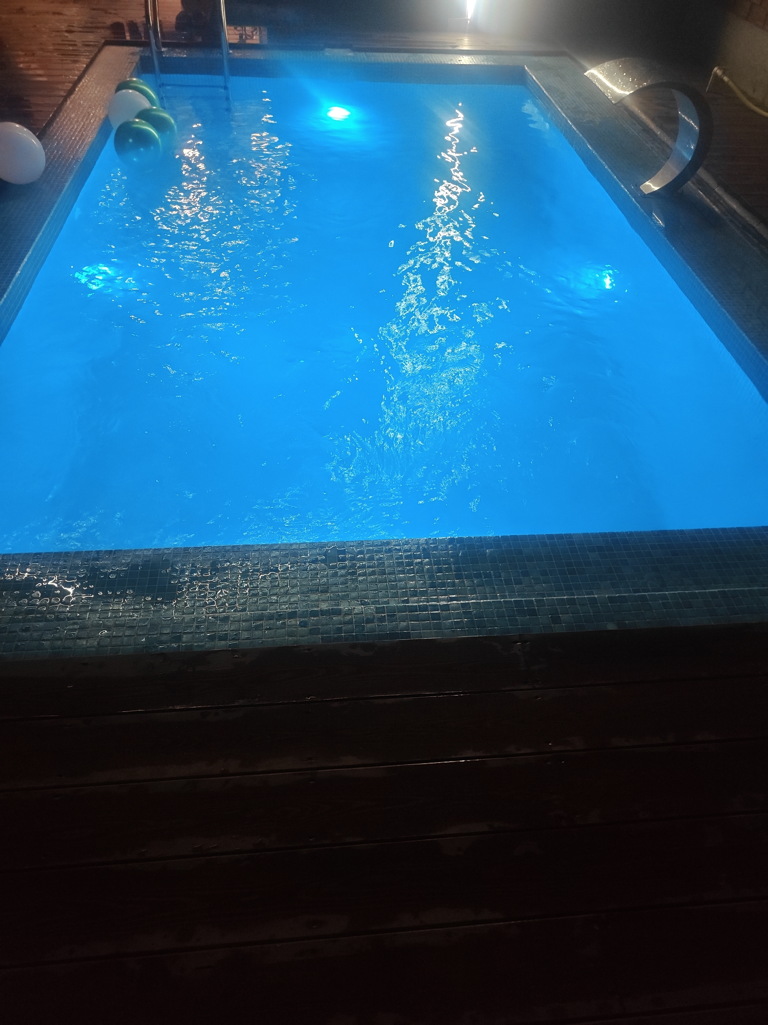 A swimming pool filled with bright blue water is lit from beneath, casting an ethereal glow. The pool is bordered by dark, tiled edges, and a wooden deck surrounds it. Several white and green balloons float on the surface of the water, suggesting a celebration. A chrome ladder is visible on the far side of the pool, and a stainless-steel handrail curves over the edge, providing support. The scene is dark, indicating it is likely nighttime. There are no people or animals visible in the image. The location is identified as Baygeldy, Kyrgyzstan.