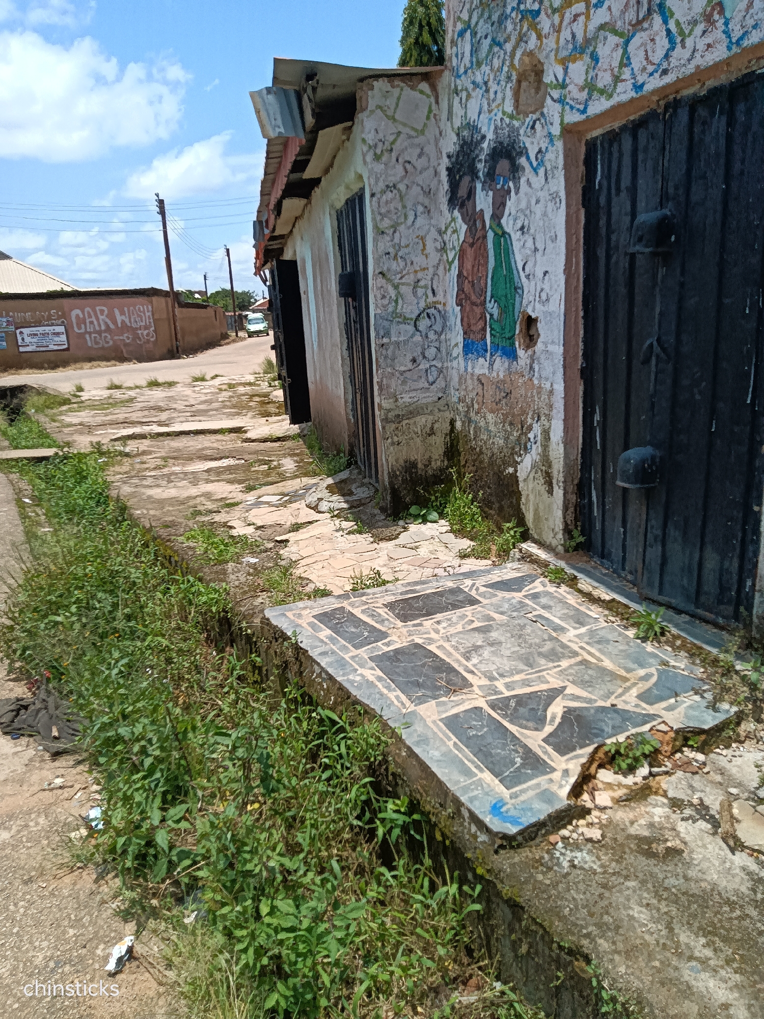 This is an eye-level, medium shot captured on a sunny day in Jos, Nigeria. The image features a street scene with a building on the right side, adorned with graffiti of two figures with prominent afros, wearing sunglasses and casual clothing. Adjacent to the graffiti are two black metal doors. The building's wall is weathered and peeling, with abstract colorful streaks of paint above the graffiti.
To the left of the building, a pathway is visible, made of uneven concrete slabs. A narrow, overgrown drainage ditch runs alongside the pathway, filled with lush green vegetation and some scattered litter. Further down the street, another building with a corrugated metal roof is visible, and a sign on its wall reads "CAR WASH." A light-colored vehicle is seen driving away in the distance. Overhead, power lines are strung between poles against a backdrop of a bright blue sky with scattered white clouds. A tiled surface, possibly a platform or step, is situated in the foreground, adjacent to the drainage ditch. The overall impression is one of a developing urban environment with signs of neglect and artistic expression.