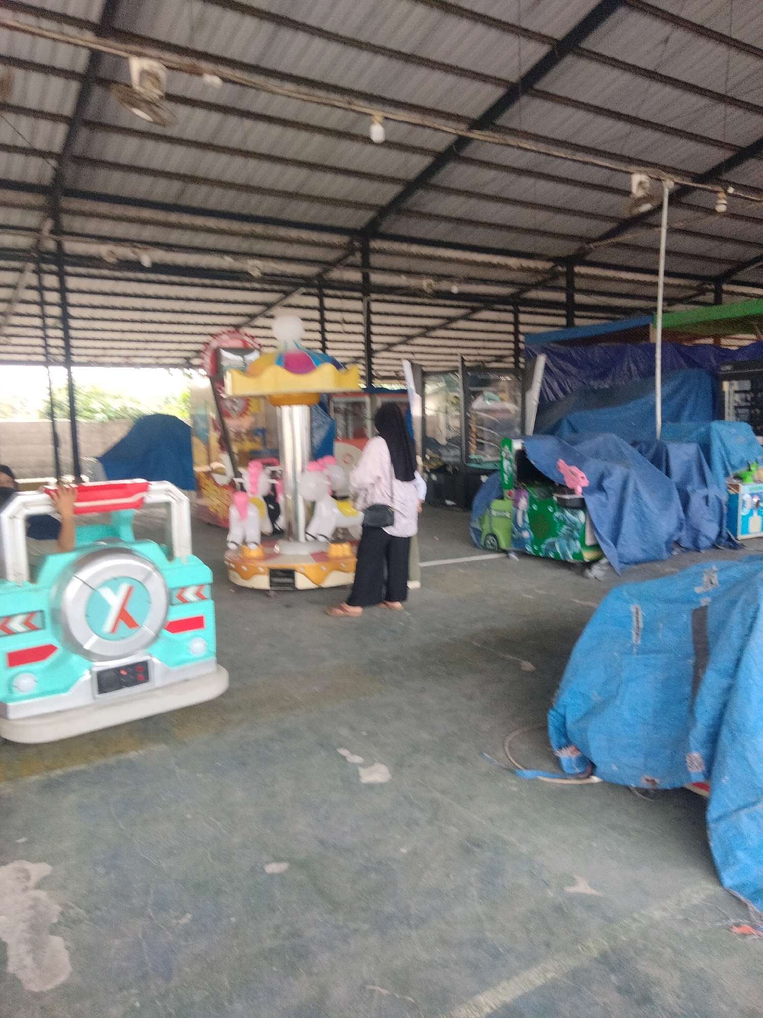 The image depicts an indoor amusement area, possibly a children's play area or a section of a larger entertainment venue, located in Sukaasih, Indonesia. The ceiling is constructed with corrugated metal roofing supported by dark metal beams, and industrial ceiling fans are visible. The floor is a rough, grey concrete surface with some lighter patches and scuff marks.
In the foreground, a mint green and white toy car ride is positioned. It has a circular silver hub with a red "X" logo on its front. A person's arm, wearing a dark blue shirt, is visible reaching out from the interior of the toy car.
Behind the toy car, a small carousel with four white horse figures is set up. A woman wearing a light pink long-sleeved shirt, black hijab, black pants, and sandals is standing next to the carousel, facing away from the camera and looking towards it. She is carrying a black bag over her right shoulder.
Further in the background, several amusement rides and structures are covered with large blue tarps. One visible ride has a yellow and colorful canopy with a yellow and blue top. To the left of the carousel, another structure with a red and yellow front is partially visible. To the right of the covered rides, a tall, narrow vertical structure with a clear plastic casing, possibly a game or attraction, is visible.
The overall lighting appears to be natural, coming from the open sides of the structure, with some artificial lighting from bulbs attached to the ceiling. The environment suggests a somewhat weathered or underutilized amusement space.