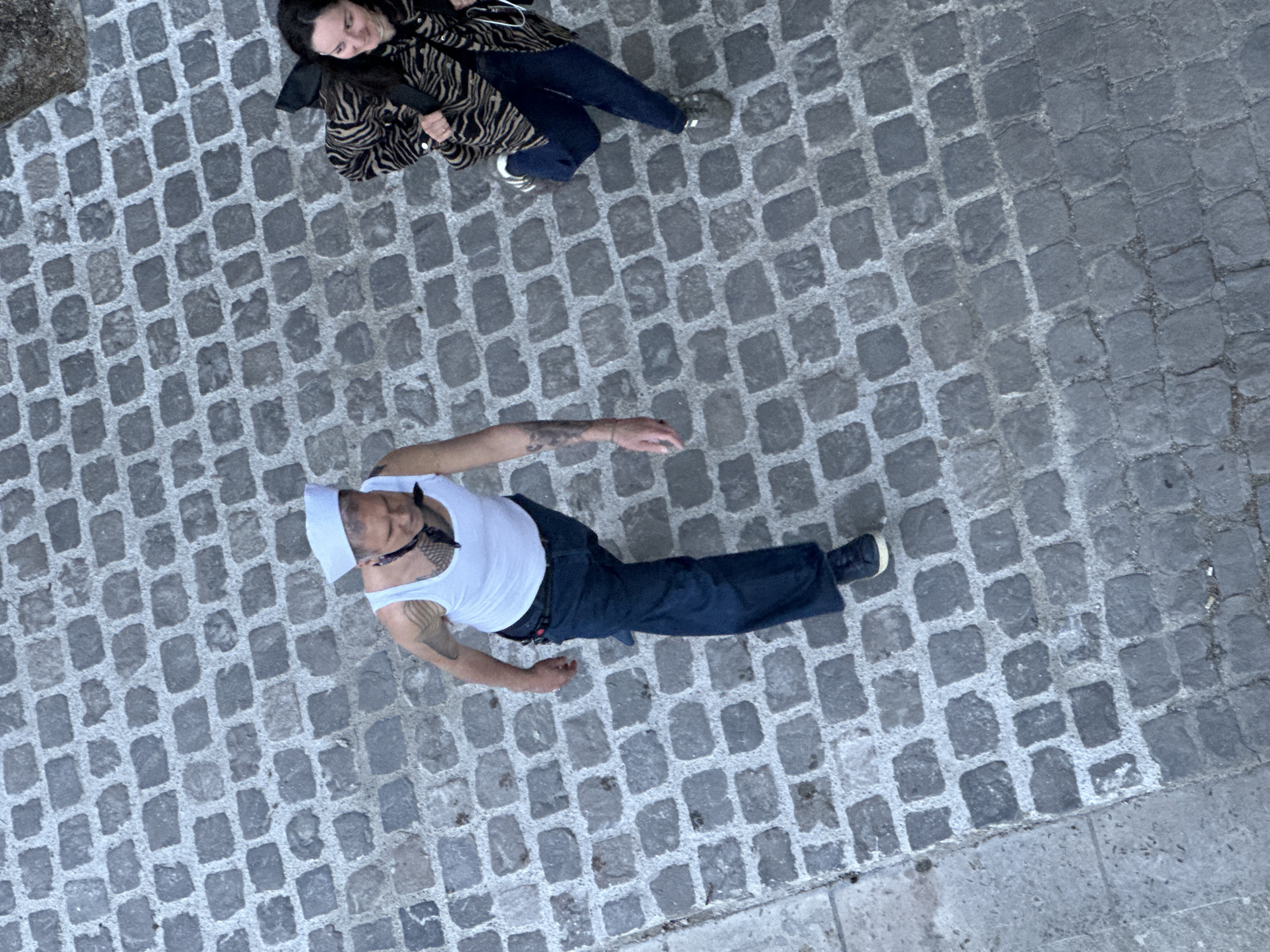 The image is a high-angle shot of a cobblestone street, likely taken from a building. Two individuals are present. One is a woman in a zebra-striped jacket and blue jeans, facing the left side of the frame. The second individual, a man wearing a white sailor hat, a white tank top, and dark blue pants, is positioned lower in the frame. He appears to have tattoos on his arms. The setting suggests an urban environment, potentially in Paris, France, due to the presence of cobblestones which are commonly seen there.