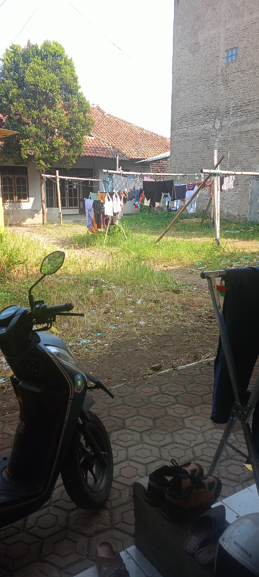 The image captures an outdoor scene in Margahayu Selatan, Indonesia, likely a residential area. In the foreground, a dark-colored scooter is parked on a patterned brick walkway. To the right, a metallic drying rack holds dark clothing. Behind the scooter and drying rack, a patch of dry grass and dirt is visible, with some scattered debris. In the mid-ground, a clothesline made of bamboo poles supports a variety of laundry items, including shirts, pants, and undergarments, which are drying in the sun. A house with a red tiled roof and wooden-framed windows is partially visible behind the clothesline. Further back, a tall, textured concrete building with a small blue-paned window stands to the right of the house. A large green tree is situated to the left of the house, casting a shadow on the ground. The sky is bright and clear, suggesting it is daytime.