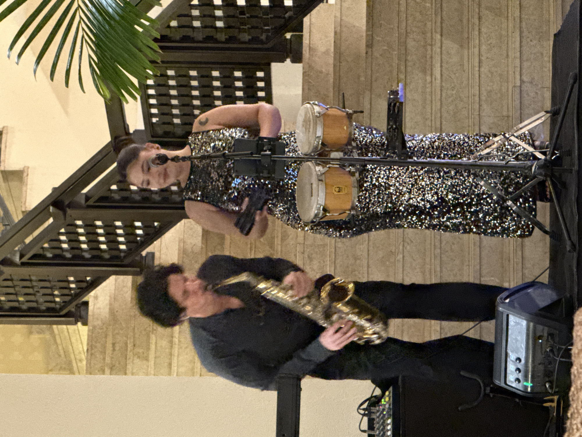 Two musicians are performing live in an indoor venue in Dubai, United Arab Emirates, set against a backdrop of a light-colored stone staircase with dark, decorative metal railings featuring a grid pattern. On the left, a male musician with dark, curly hair wears a dark long-sleeved shirt and black trousers, actively playing a gold-colored saxophone. His gaze is directed downwards at the instrument. To his right, a female musician with dark hair pulled back into a bun stands behind a microphone stand and a set of light-wood bongos. She wears a sleeveless, floor-length gown adorned with shimmering black and silver sequins and has a crescent moon tattoo on her left upper arm. She holds a dark, rectangular object in her left hand and looks slightly to her left with a subtle smile. A black vocal microphone is positioned on a stand in front of her, which also supports a small electronic device holder above the bongos. On the left side of the scene, a small audio mixer with multiple knobs sits atop a larger black speaker or equipment case. On the floor in the foreground, a black wedge-shaped monitor speaker with a green indicator light is visible. A large green palm frond is present on the far right, partially obscuring the background. The musicians are standing on a dark, raised platform or carpeted area, under warm, artificial lighting.
