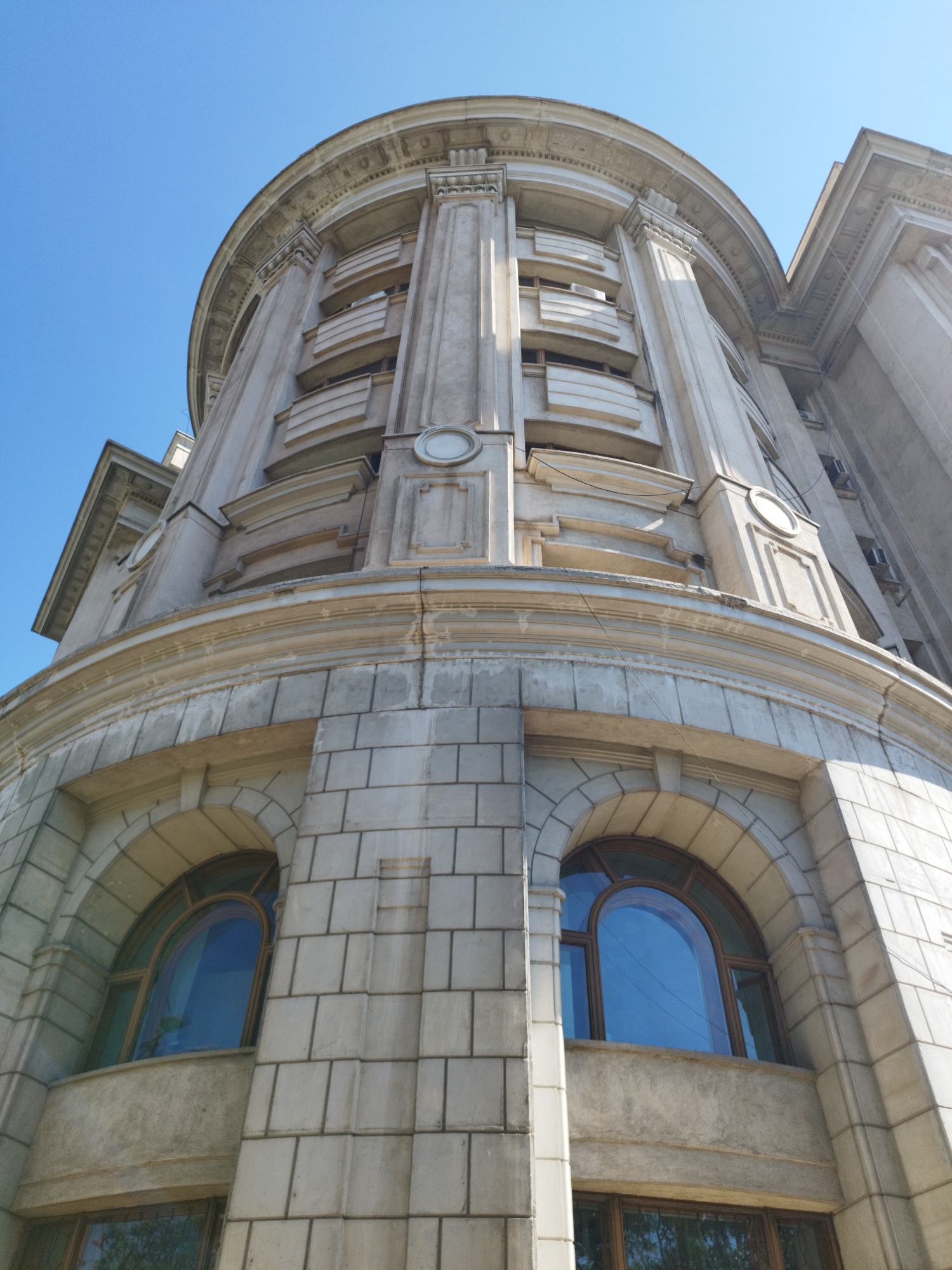 The image shows a low-angle, upward view of a grand, historic building in Bucharest, Romania, under a clear blue sky. The architecture is characterized by its imposing, curved facade, adorned with classical elements such as columns, decorative moldings, and arched windows. The building appears to be constructed of light-colored stone or concrete, showing some signs of age and weathering.
The facade features multiple stories, with recessed balconies or window areas on the upper levels, distinguished by horizontal banding and protruding architectural details. Circular decorative elements, possibly medallions or oculi, are visible on some sections of the wall. The windows themselves are large and have arched tops, with the panes reflecting the bright sky. The lower portion of the building shows large, rectangular stone blocks, creating a textured and robust base.
The overall impression is one of architectural grandeur and historical significance, with the upward perspective emphasizing the building's scale and majesty. No people or specific contemporary objects are discernible in the image. The scene is purely focused on the building and its immediate surroundings, which are implied to be an urban environment due to the architectural style and presence of what appear to be air conditioning units on some of the upper floors, though these are not clearly detailed.