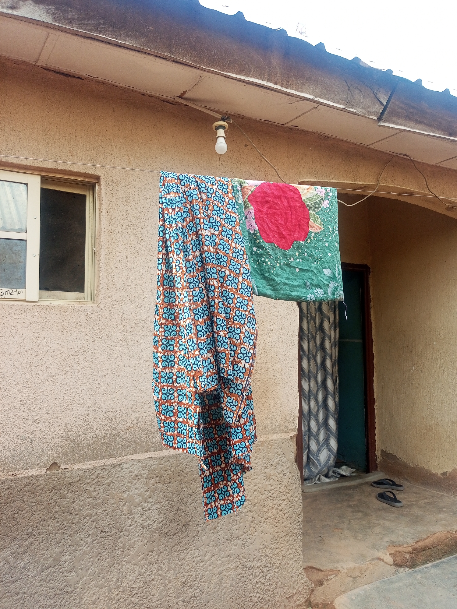This is an outdoor shot of laundry drying on a clothesline in Jalingo, Nigeria. Two pieces of fabric are hanging, one is a colorful patterned cloth with blue and orange swirls, and the other is a green fabric with a large red floral design. Behind the laundry, a dark green door is visible, partially obscured by a patterned curtain. A pair of dark-colored flip-flops rests on the concrete floor in front of the door. The building has a light brown, textured exterior wall, and a section of its roof with a white corrugated metal overhang is visible at the top of the frame. A single light bulb hangs from a wire. The scene appears to be set during the daytime, under potentially overcast or hazy skies, as there are no strong shadows. The overall impression is a simple, everyday scene in a residential area. Text is visible on the wall to the left of the window, appearing to read "am2-101".