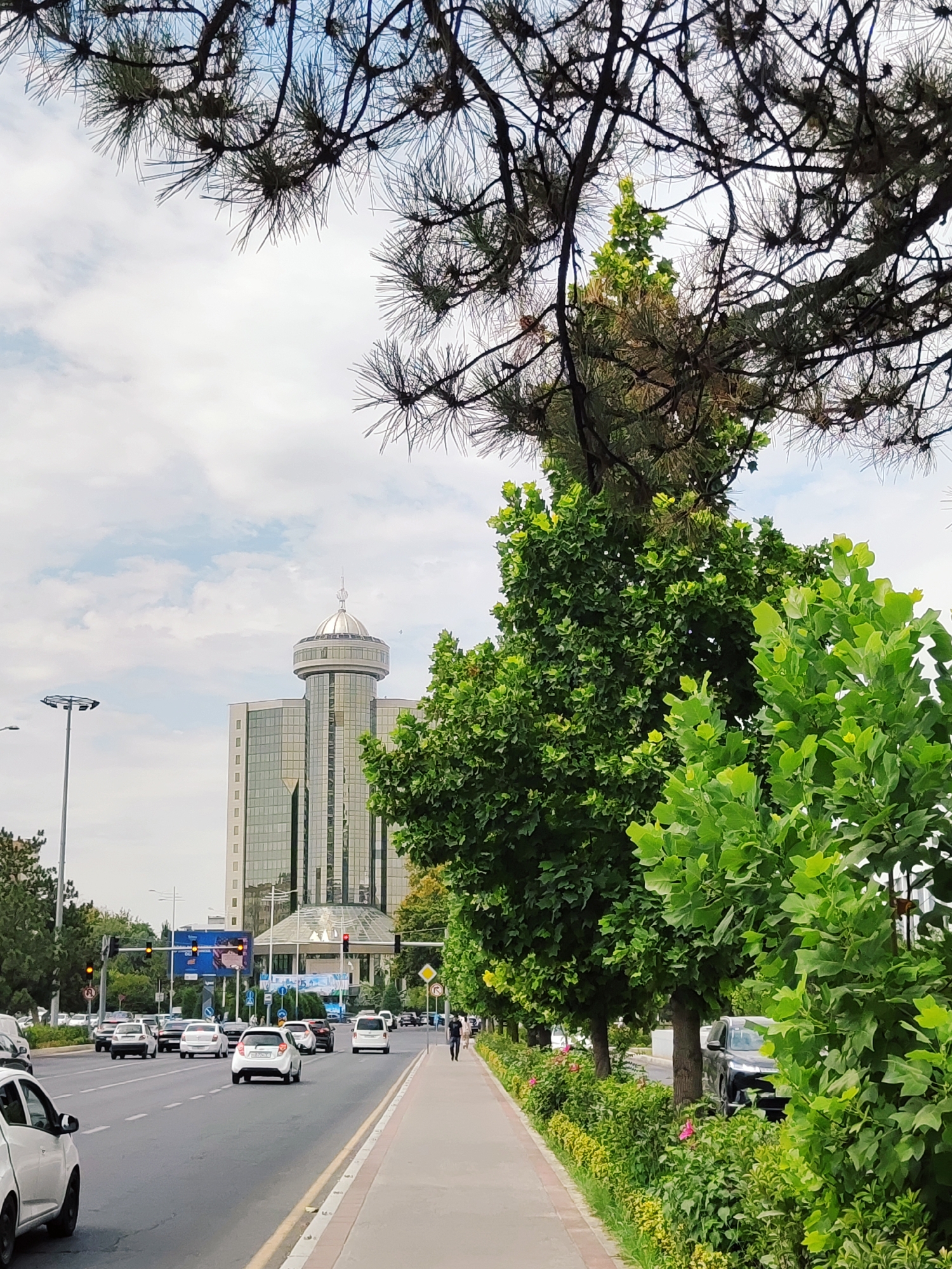 The image depicts a street scene in Tashkent, Uzbekistan, with a modern glass building in the background. In the foreground, a busy road with multiple lanes of traffic is visible, with several cars driving in the same direction. A pedestrian walkway runs alongside the road, lined with trees and green foliage. A man is walking on the sidewalk. The sky is partly cloudy. The overall impression is that of an urban environment with greenery interspersed with modern architecture and infrastructure.