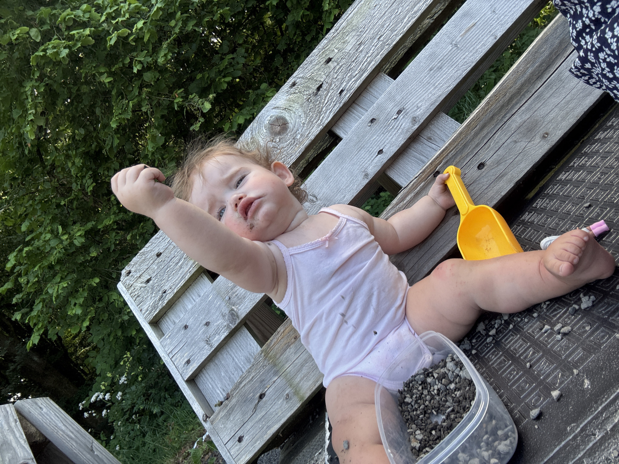 A high-angle medium shot captures a young child, likely a toddler, lying on a weathered wooden pallet bench. The child is wearing a light pink onesie that is smudged with dirt. Their right arm is extended upwards, with their hand open as if reaching for something. Their face shows a slightly grumpy or tired expression, with dirt smudges around their mouth.
Beside the child, a bright yellow toy shovel rests on the bench. A clear plastic container filled with small grey pebbles is also visible. The background consists of lush green foliage, suggesting an outdoor setting. The lighting indicates it is daytime, likely outdoors in a garden or natural area. The overall scene depicts a child playing in the dirt. There is no visible text in the image.