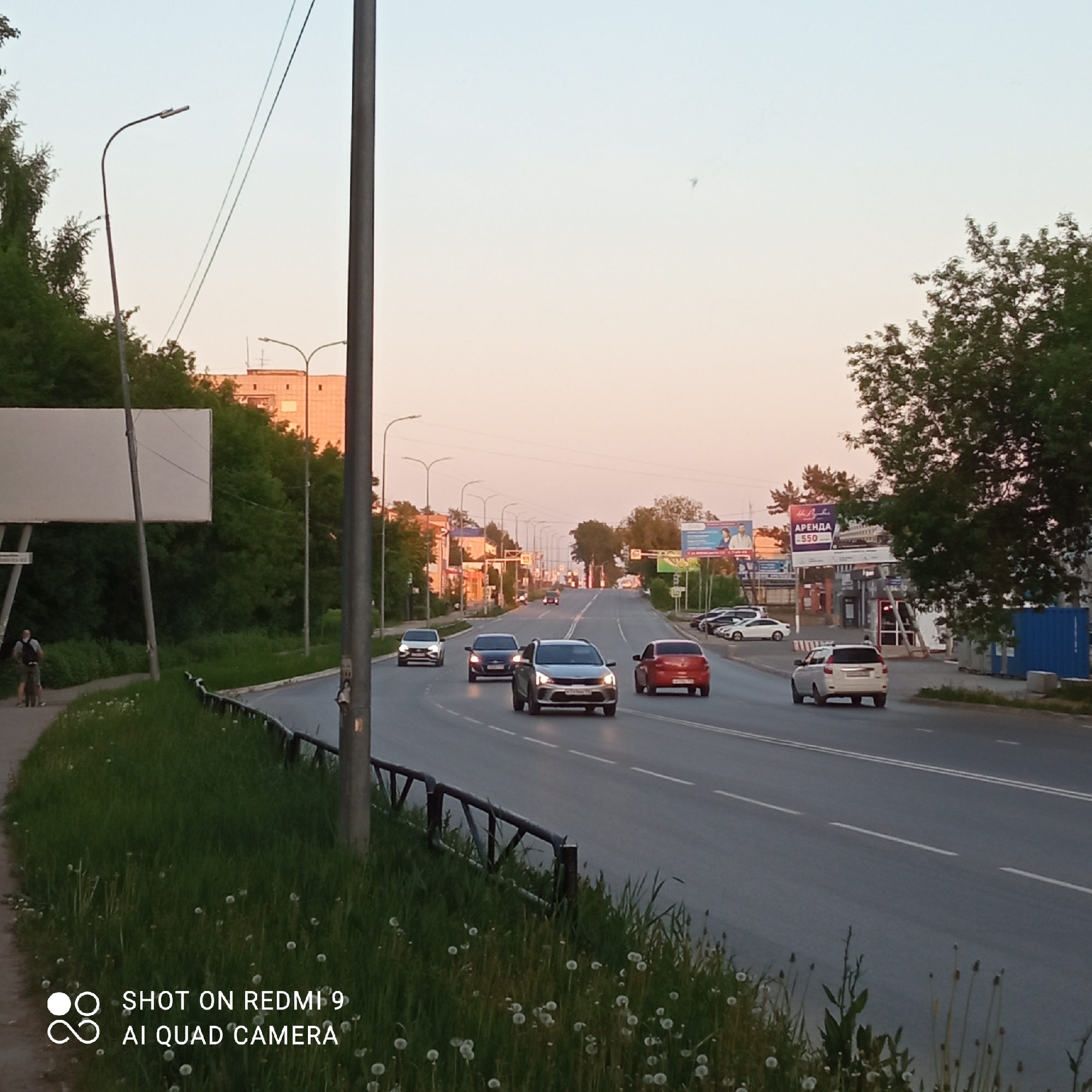 The image shows a street scene in Chaikovsky, Russia. Cars are traveling on a multi-lane road that curves slightly. The sky is a pale pink, suggesting either sunrise or sunset.
On the left, there's a grassy area with dandelions and a sidewalk. Two individuals, one possibly on a bicycle, are walking along the sidewalk next to a large, blank billboard. A metal guardrail separates the sidewalk from the road.
On the right side of the road, there's a gas station with signs in Russian, including one advertising "ARENDA" (rent) for 550. Other parked cars are visible near the gas station. Trees and buildings line the background. Streetlights are present on both sides of the road.
The bottom of the image contains the text "SHOT ON REDMI 9 AI QUAD CAMERA."