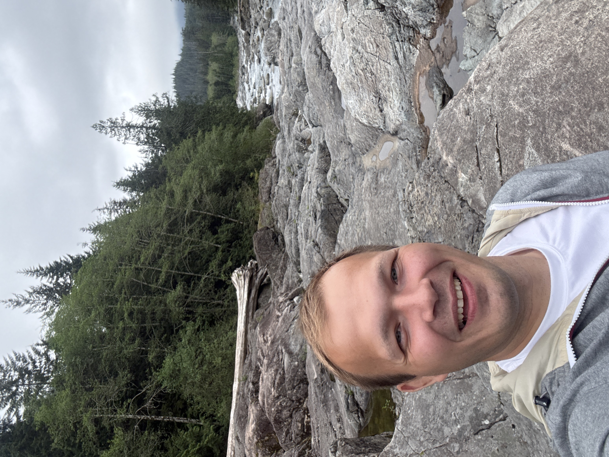 A selfie of a person with light skin and short blonde hair, wearing a white t-shirt and a light-colored jacket with a gray hoodie, against a backdrop of a rocky area with trees and a body of water. The location is within Area C, Long Beach, Canada, identifiable by the unique rock formations, vegetation, and water flow characteristics of the area. A dead tree stump can be seen within the rocky area. The sky is overcast.