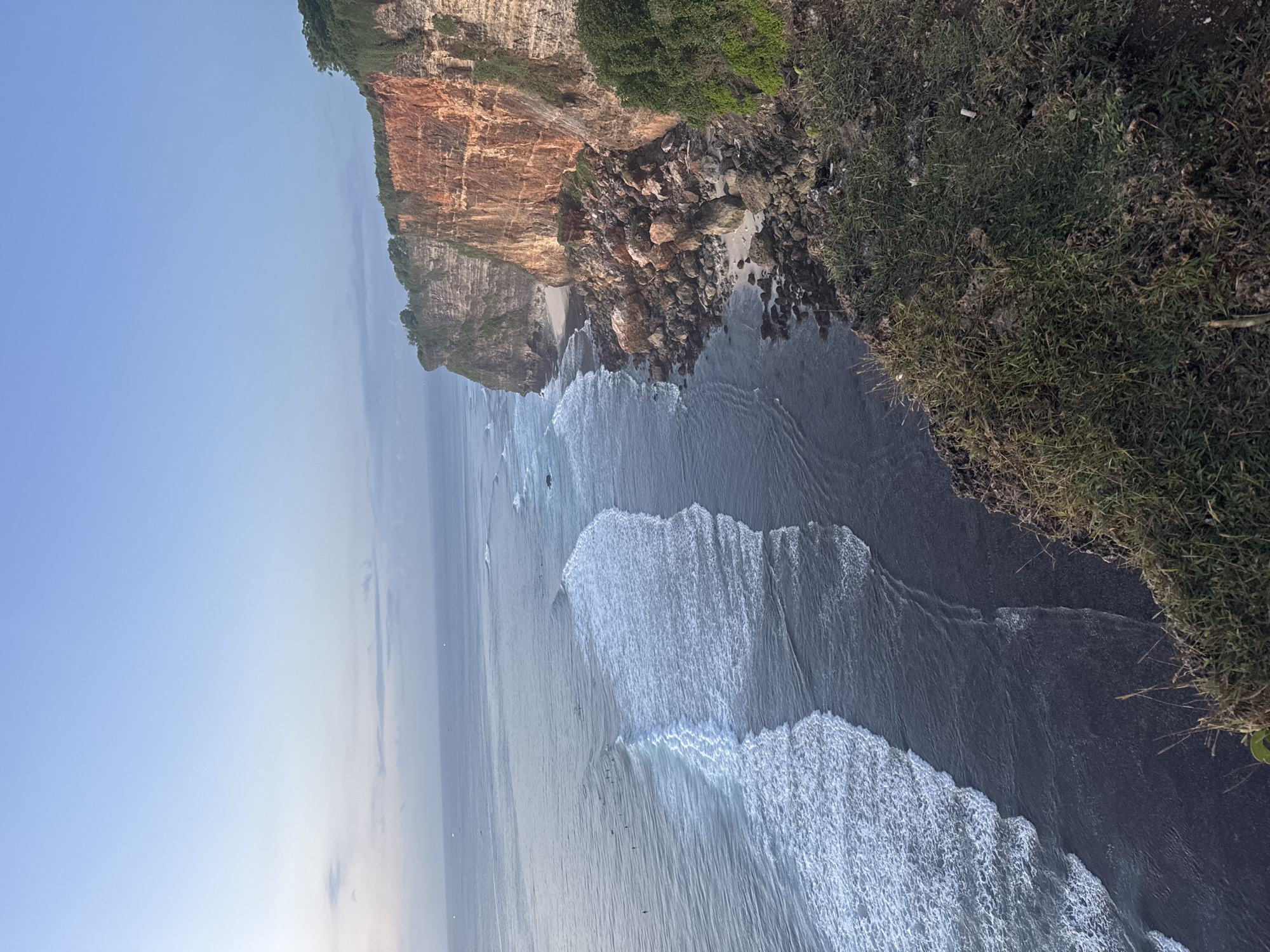 The image depicts a coastline scene with a high, rocky cliff on the right side. The cliff face shows a mix of orange and brown tones, with vegetation at the top. Below the cliff, there's a rocky area with some sand visible. The ocean stretches out, with visible waves crashing onto the shore. The sky is a light blue color, fading into a lighter hue closer to the horizon. The location is Unknown, Unknown. There is no sign of human activity.