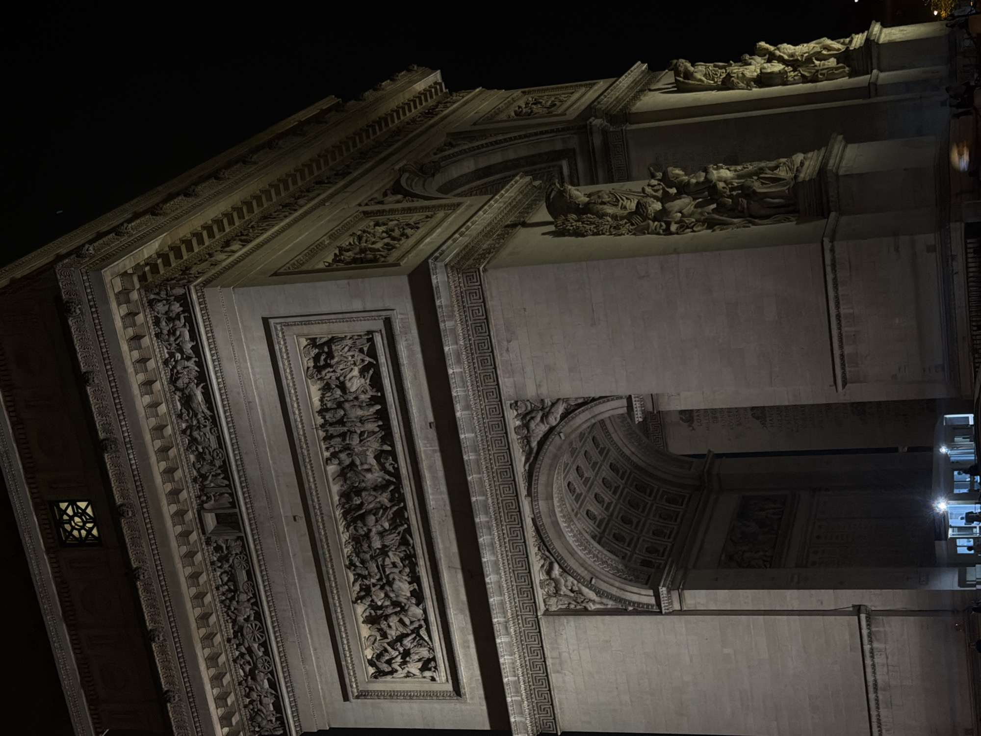The image shows a night-time view of the Arc de Triomphe in Paris, France. The arch is brightly lit, highlighting its detailed carvings and sculptures. The sky is dark and featureless. People are visible inside and underneath the arch.