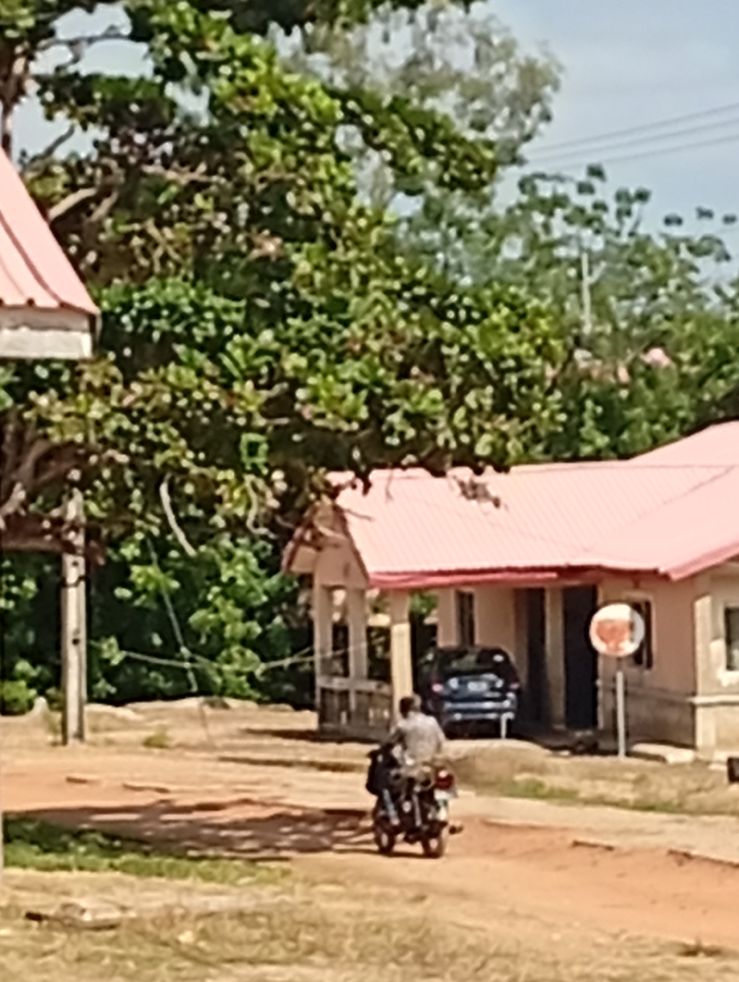 A person on a motorcycle is riding on a dirt road in front of a pink-roofed building. The person is wearing a light-colored shirt and dark pants. The motorcycle is black with a visible exhaust pipe. In the background, there are green trees and a blue car. The building appears to be a small structure with an open entrance and a round sign on a pole. The ground is covered with dirt and some patches of grass. The sky is partly cloudy.