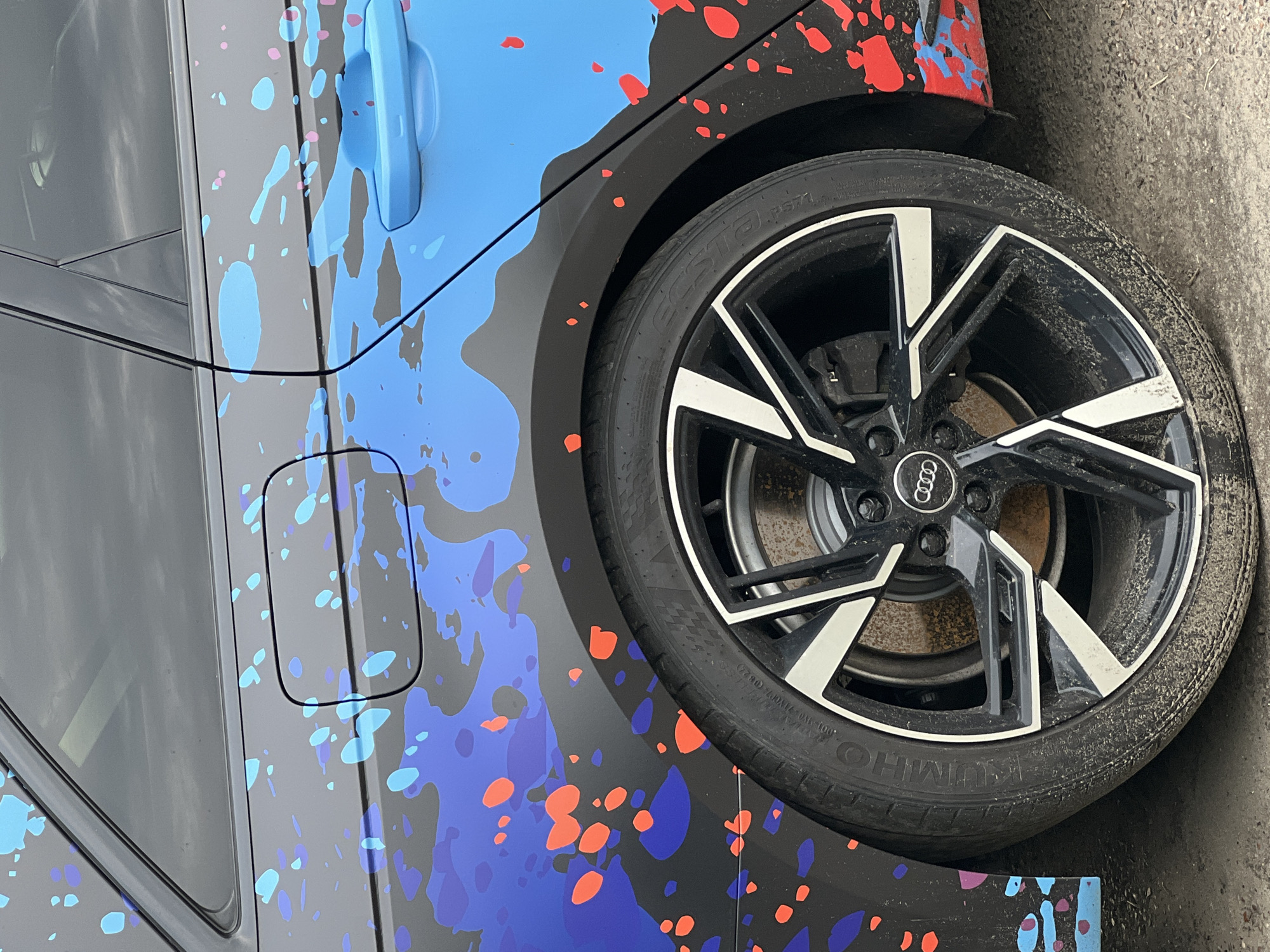 The image shows a close-up of the rear wheel and fender of a dark-colored Audi with a custom graphic wrap. The wrap features a chaotic design of splattered colors, including vibrant blues, purples, and reddish-orange dots, overlaid on the car's body panels. The fuel cap is visible on the rear quarter panel, and the door handle, also wrapped in the graphic, is partially visible. The car's wheel is a black multi-spoke alloy rim with silver accents, and the tire is dirty, suggesting it has been driven on a rough surface. The brake rotor and caliper are visible behind the spokes. The background is slightly out of focus but appears to be a paved surface, possibly a road or parking lot. The image does not provide any specific information that would allow for its location to be identified as Krasnodar, Russia. There are no people or any actions depicted in the scene.