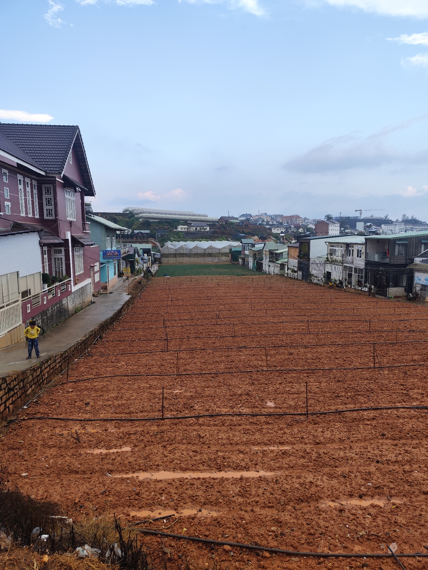 The image captures a scene in Lang Biang - Đà Lạt, Vietnam. A large, recently tilled field of reddish-brown soil dominates the foreground. Horizontal black irrigation pipes run across the field, supported by vertical stakes. In the background, houses and buildings are visible, typical of a densely populated area. Greenhouses are seen on a distant hill. To the left, a multi-story building with a red exterior and white trim is partially visible. A small boy wearing a yellow shirt and blue pants stands on a low stone wall next to the building. A narrow paved street separates the building and the field. The sky is overcast with scattered clouds. Debris is scattered at the bottom of the frame.