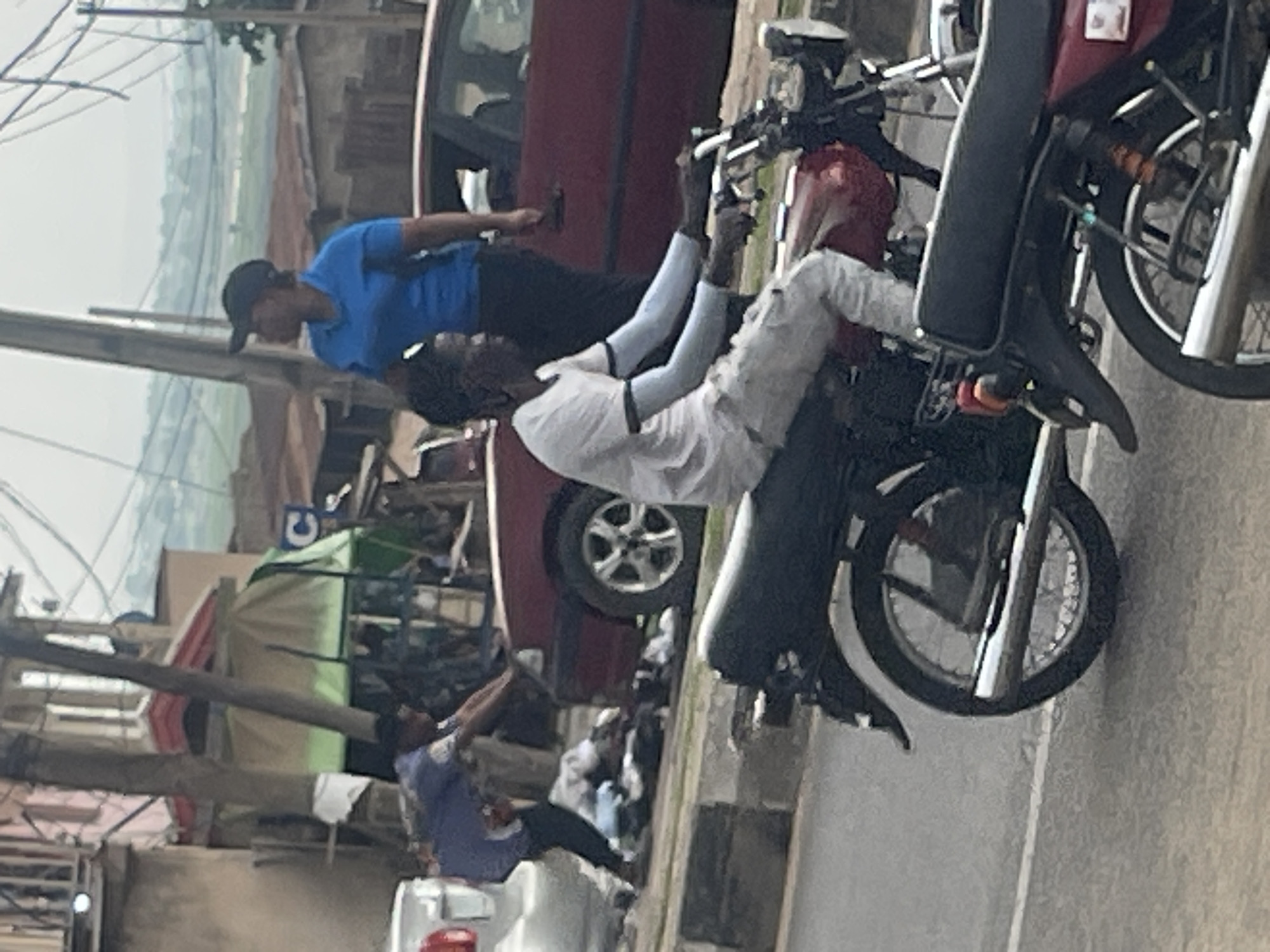 An overhead view captures a street scene in an urban environment, likely in a developing country. A person is seen on a red motorcycle in the foreground, wearing a white t-shirt and light-colored pants, with arm sleeves. Behind them, a maroon car is parked or moving slowly on the road. To the left of the car, another person is partially visible, appearing to be interacting with a pile of clothing on the ground near the curb. Further in the background, a woman in a blue shirt and a black cap is standing, and several buildings with corrugated metal roofs are visible, along with utility poles and power lines overhead. The overall impression is one of a busy street with various activities taking place.