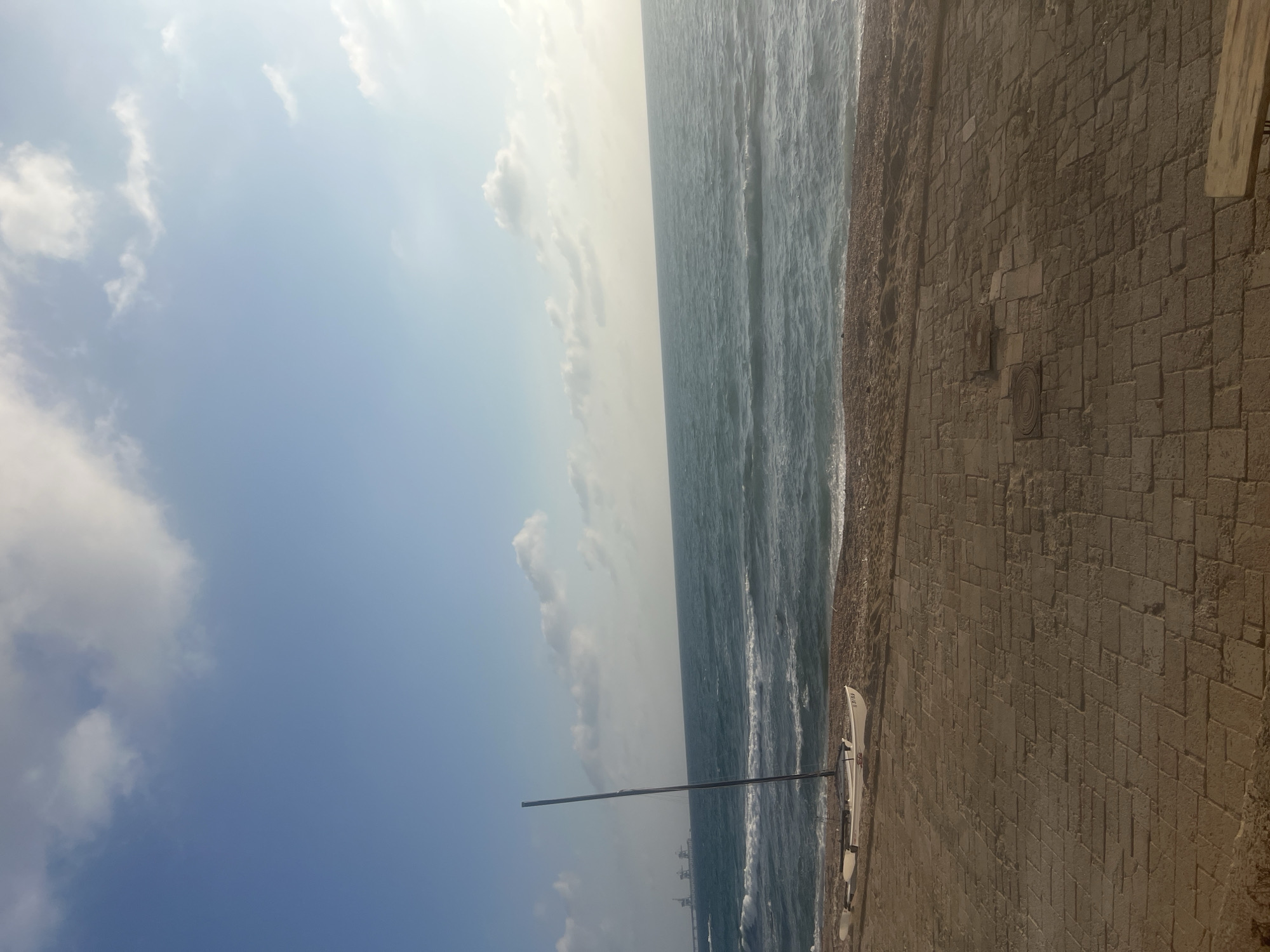 This media file depicts a seaside scene with a paved walkway bordering the ocean. The walkway is constructed of large, rectangular stone blocks laid in a herringbone pattern. To the left of the walkway, the sea is visible, with moderate waves breaking near the shore. A small, white sailboat with a black mast is resting on the sand near the water's edge. In the distance, several industrial structures, possibly cranes or buildings, are faintly visible against the hazy horizon.
The sky is a light blue with scattered, fluffy white clouds. The lighting suggests it is daytime, likely morning or late afternoon, with a soft, diffused sunlight. The overall mood is tranquil and serene.
The image is oriented vertically, making it appear as though the viewer is looking up from a low angle. Some text is visible on the side of the sailboat, but it is too small and blurry to be deciphered. The setting is consistent with a coastal area in Hof HaCarmel Regional Council, Israel, which features paved promenades and views of the Mediterranean Sea.