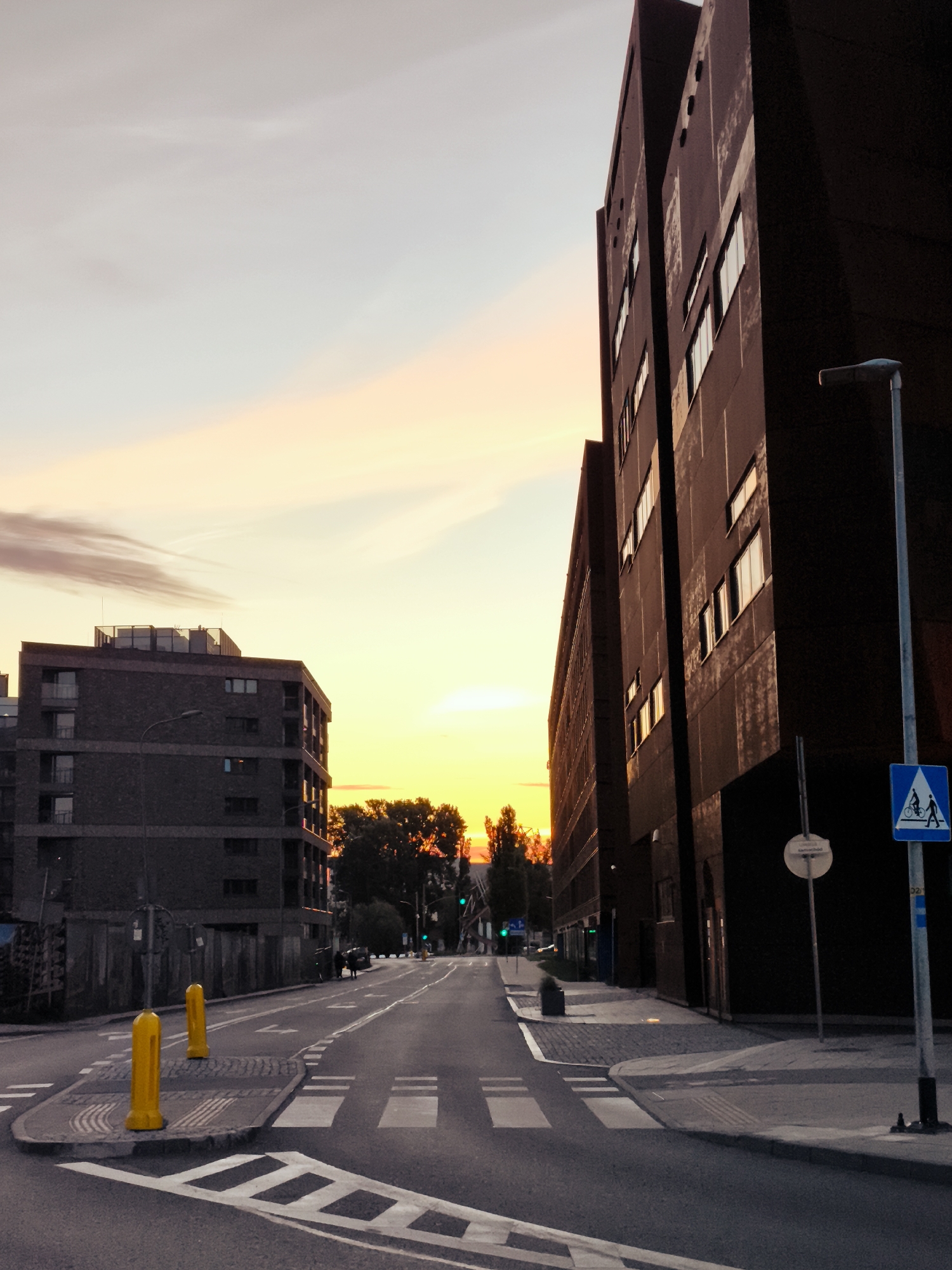 The image depicts a street scene in the city of Gdańsk, Poland, during sunset. On the right, a large, dark, modern building with multiple windows dominates the foreground. A lamppost with a blue and white pedestrian crossing sign is visible in front of this building. On the left, a smaller, multi-story building with a brick facade and balconies is situated further down the street. The street itself is paved and features a pedestrian crossing with white markings. Two bright yellow bollards stand on a raised section of pavement near the pedestrian crossing. The sky above is filled with soft, warm hues of orange and yellow, characteristic of a sunset, with some wispy clouds. Trees are visible in the middle ground, silhouetted against the bright sky. The overall atmosphere is calm and urban.