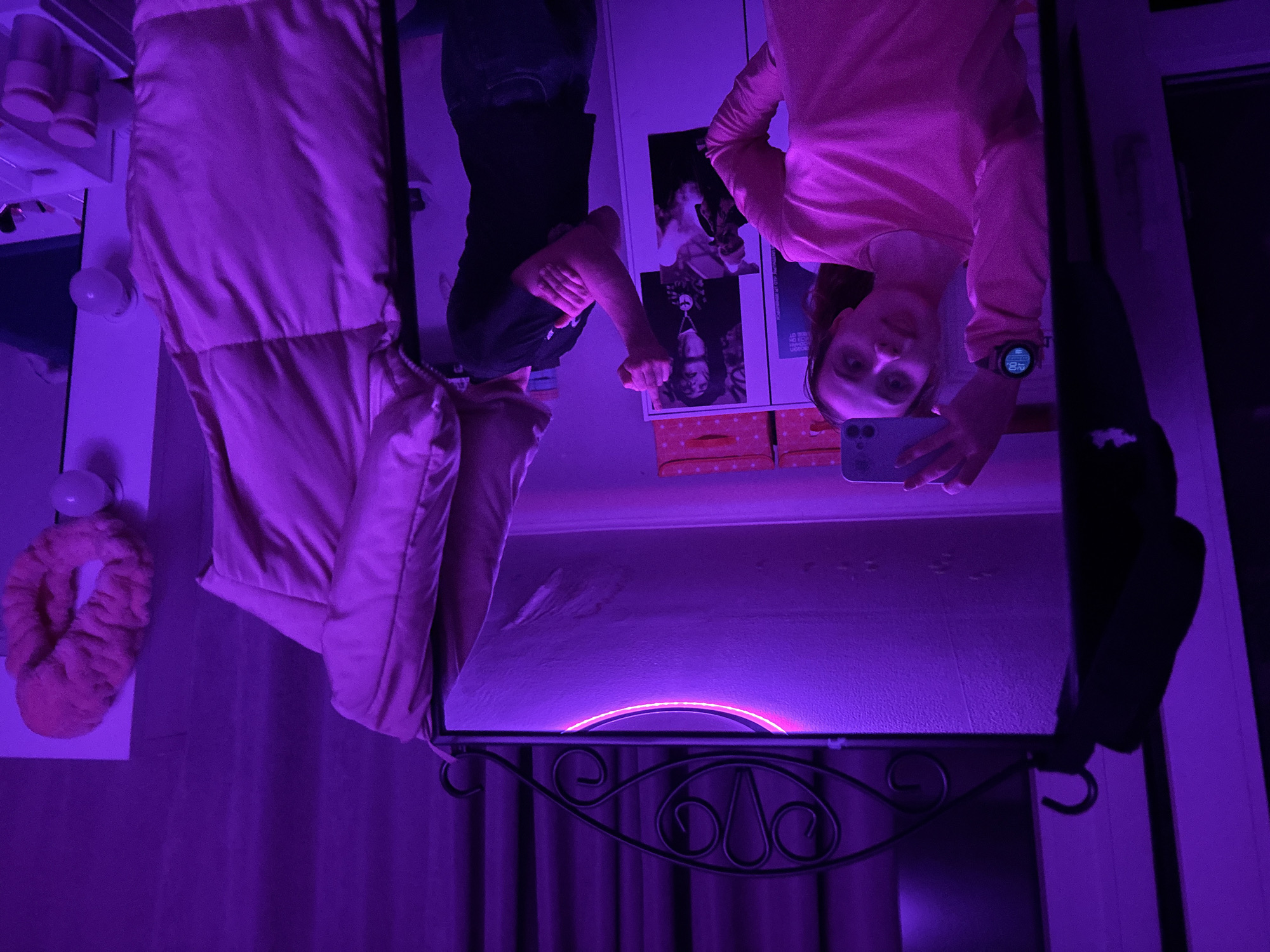 This image, set in Прикамский, Russia, captures a mirror selfie taken in a room bathed in vibrant purple and pink light, giving it a distinct, atmospheric mood. The entire image is rotated 180 degrees.
The main subject is a young woman, seen in her reflection, who is taking the photo with a light-colored smartphone (likely an iPhone) held up towards the mirror. She is wearing a long-sleeved pink top and has a dark smartwatch on her left wrist. Her expression is neutral to slightly smiling. On the smartwatch, the digital time "11:02" is visible, along with other small icons and possibly the day "ПТ" (Fri).
The scene is an indoor setting, likely a bedroom or dressing area, indicated by the presence of a vanity-style mirror and personal items. The mirror is framed at the bottom with an ornate, dark metal scrollwork design. Along what appears to be the left edge of the mirror (in the inverted image), several round light bulbs are visible, contributing to the bright, colorful illumination.
Reflected in the background behind the woman are posters or pictures on a wall; one features a person with a peace sign necklace and partially visible text, another appears to be a portrait, possibly of Tupac Shakur. Below these reflections, two polka-dotted orange or pink boxes are stacked. To the left of the mirror, a pink quilted jacket or coat hangs. Further left, a fluffy pink headband or slipper-like item is hanging on a wall. Dark purple curtains are visible in the foreground/background, adding to the room's color scheme. Another person, or at least their torso and arm wearing dark clothing, is partially reflected near the top-center of the mirror, seemingly pointing or gesturing with a closed fist. An intense, arched light source, emitting bright purple light, is reflected near the top edge of the mirror, likely a ring light or LED strip.
The predominant artificial lighting suggests the photo was taken in the evening or at night.