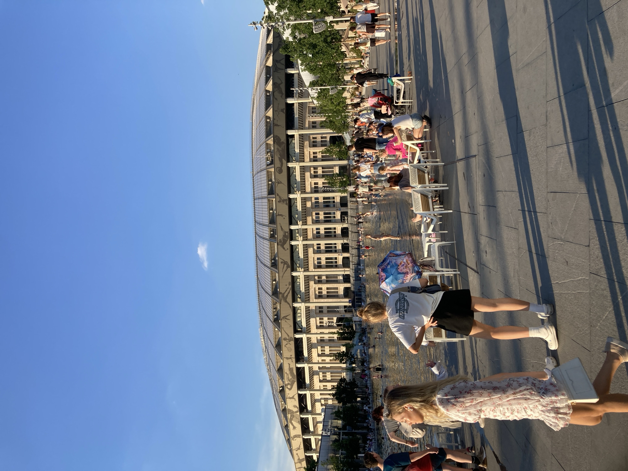 The image depicts a sunny day at Luzhniki Stadium in Moscow, Russia, with a large crowd gathered around a shallow water feature. In the foreground, a young woman in a floral dress walks past other individuals relaxing in chairs. The background shows the impressive architecture of Luzhniki Stadium, a prominent landmark in the city. Many people are wading and playing in the water, while others are seated or standing nearby. The scene is characteristic of a public gathering space on a warm day.