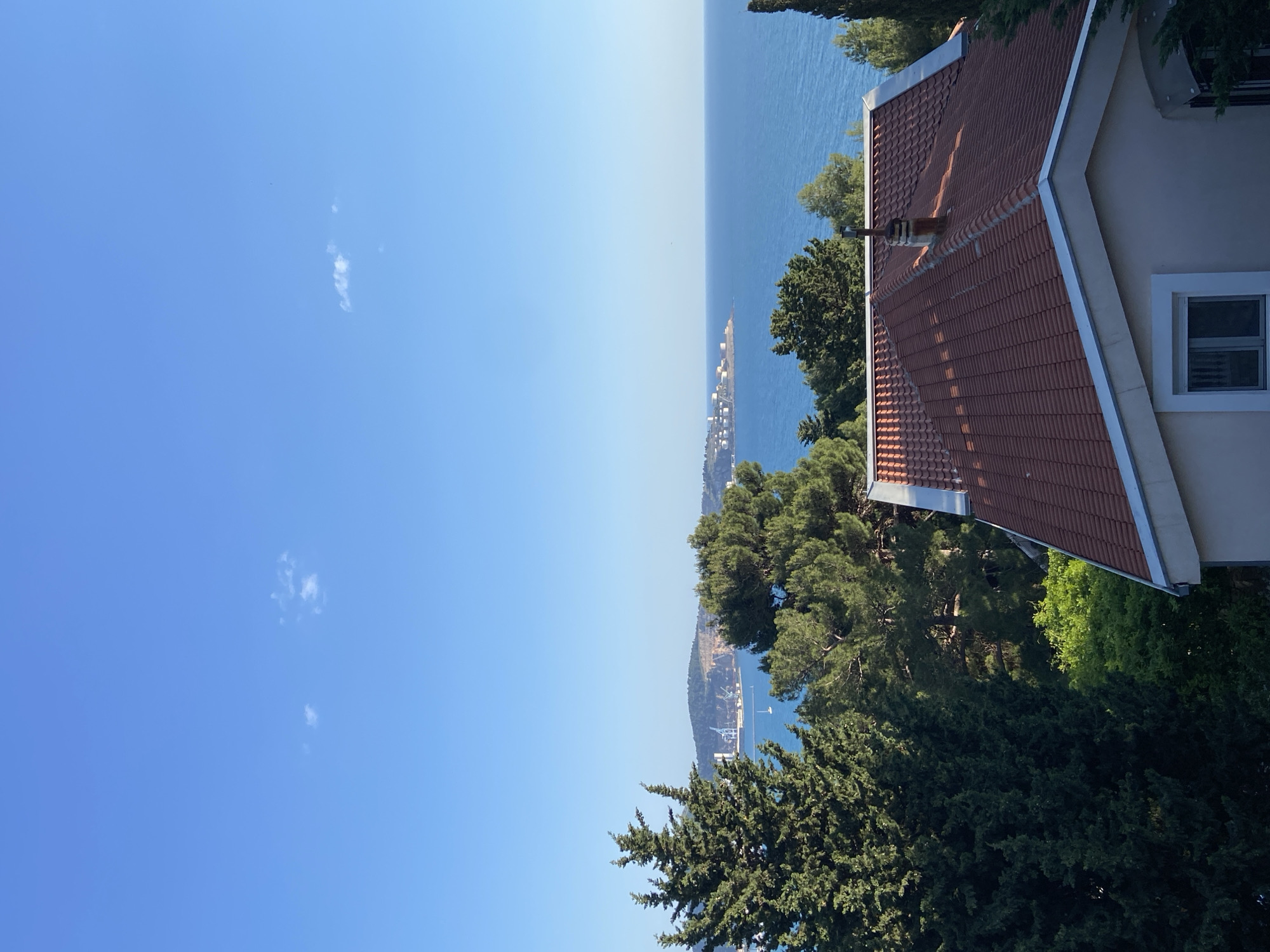 The image presents a seaside view in Šušanj, Montenegro. To the right is the corner of a building, featuring a red-tiled roof and a window. Below the building are various trees. The background is dominated by a strip of blue water and sky, separated by a distant horizon. In the middle distance, a coastline with a structure is visible. The sky is clear, with a few small clouds.