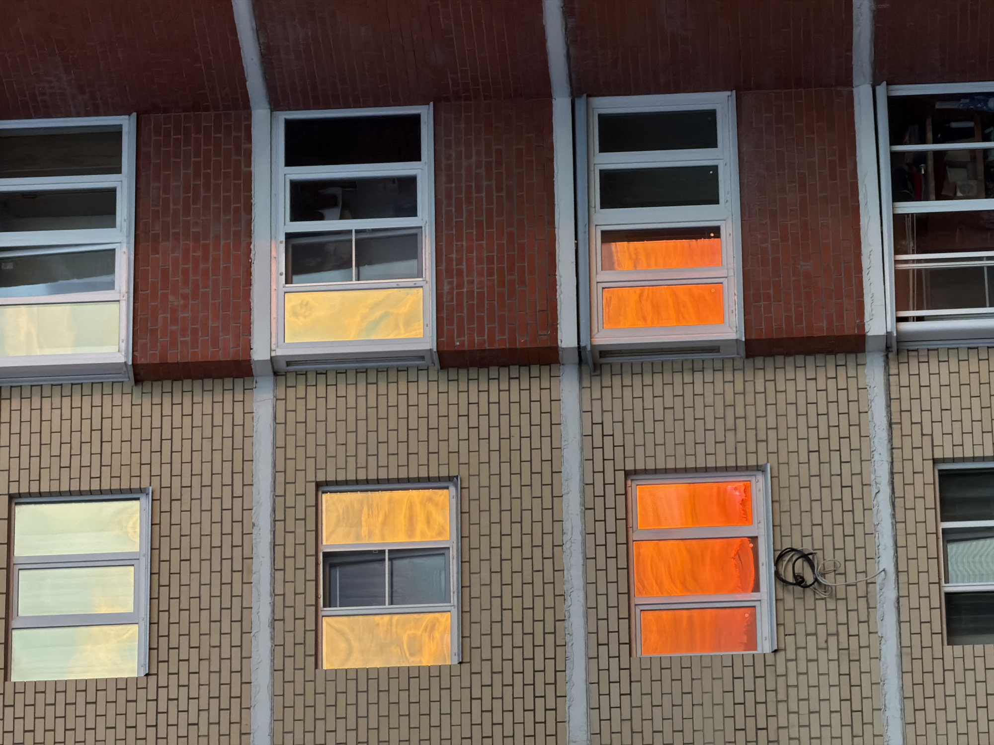 A close-up, eye-level shot captures a section of a building facade dominated by rectangular windows and brickwork, likely at either sunrise or sunset due to the warm lighting. The building is visibly divided into two horizontal sections by differing brick colors.
The upper section features dark reddish-brown bricks, extending to an overhang above the windows. This section contains four windows with white frames. The leftmost window has a subtle light yellow reflection. The next window shows a vibrant golden yellow reflection on its lower two panels. The third window displays intense orange-red reflections on its lower two panels. The rightmost window on the top row offers a view into an interior room, where various objects are dimly visible, along with some subtle reflections.
The lower section of the building is constructed with light beige bricks. It features three visible windows, also with white frames. The leftmost window in this section reflects a pale yellow or light green hue across its three panels. The middle window shows a bright golden orange reflection on its upper and lower panels, with a smaller, darker interior window visible in its center. The rightmost window in the lower section exhibits vivid orange-red reflections across all its panels. To the right of this window, a bundle of black and white electrical wires or cables is coiled and affixed to the beige brick wall.
The distinct golden and orange-red hues reflected in the windows suggest a low sun angle and clear skies. There are no people or discernible text visible in the image.