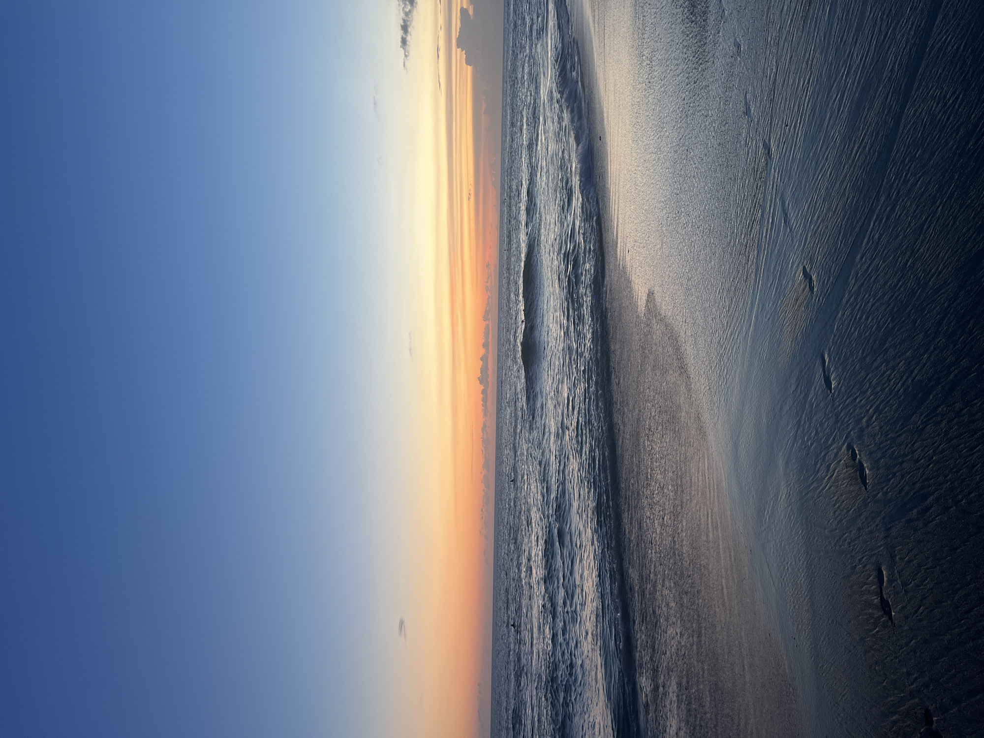 The image captures a vertical view of a beach scene, likely taken at sunrise or sunset. The left side of the frame is dominated by a clear, gradient sky transitioning from a deep blue to a soft orange at the horizon line. The right side depicts the shoreline, with gentle waves washing onto the sand. The wet sand reflects the colors of the sky, creating a mirror-like effect. No people are visible in the frame. Based on the location provided, this scene could be a beach in North Kuta, Indonesia.