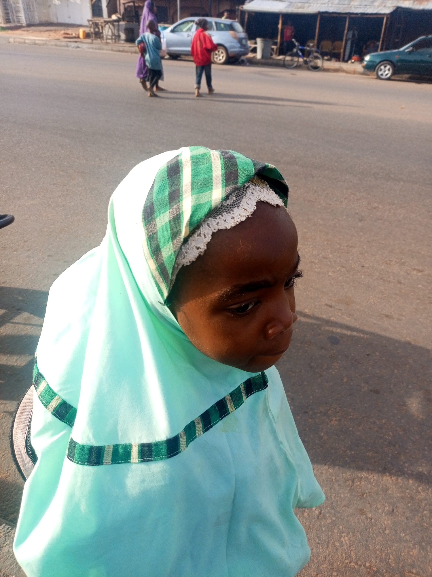 A young girl wearing a light blue hijab with green and white checkered trim stands on an asphalt road. The hijab also has white lace near the hairline. She is facing away from the camera, looking to the right. In the background, there are several people, cars, and buildings. It is daytime. The image appears to be taken in an urban setting, likely the city of Gombe, Nigeria.
