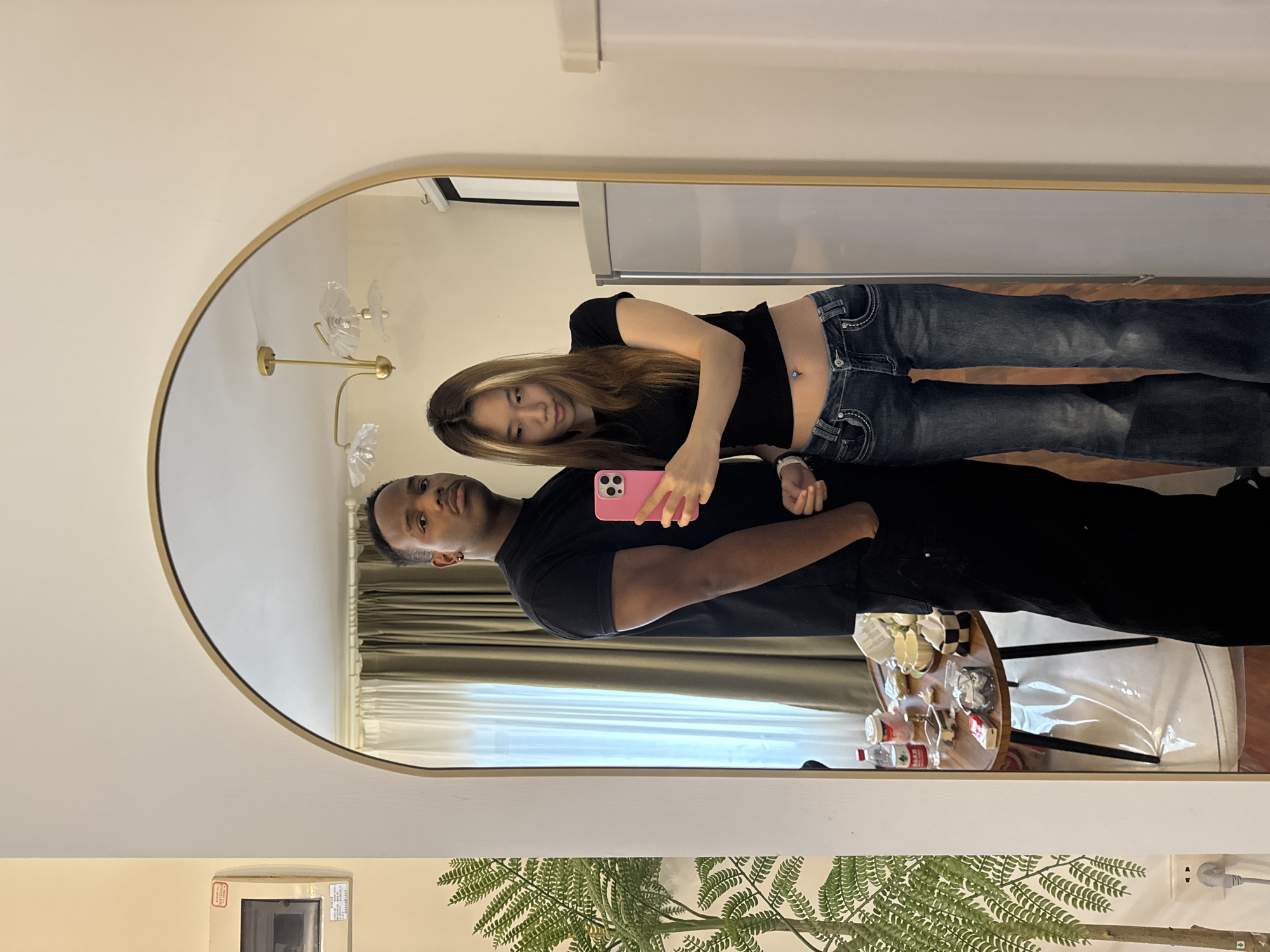 A couple poses for a selfie in front of an oval mirror with a gold frame. The mirror reflects a scene with a beige wall, a sheer curtain covering a window, and a small round table holding bottles and decorative items. The woman has long brown hair and wears a black top and blue jeans. The man wears a black T-shirt and holds a pink smartphone. The location is possibly a residential interior. The presence of a power outlet with a foreign plug suggests the location may be in the Yuzhong District, China, but this cannot be confirmed based solely on this image.