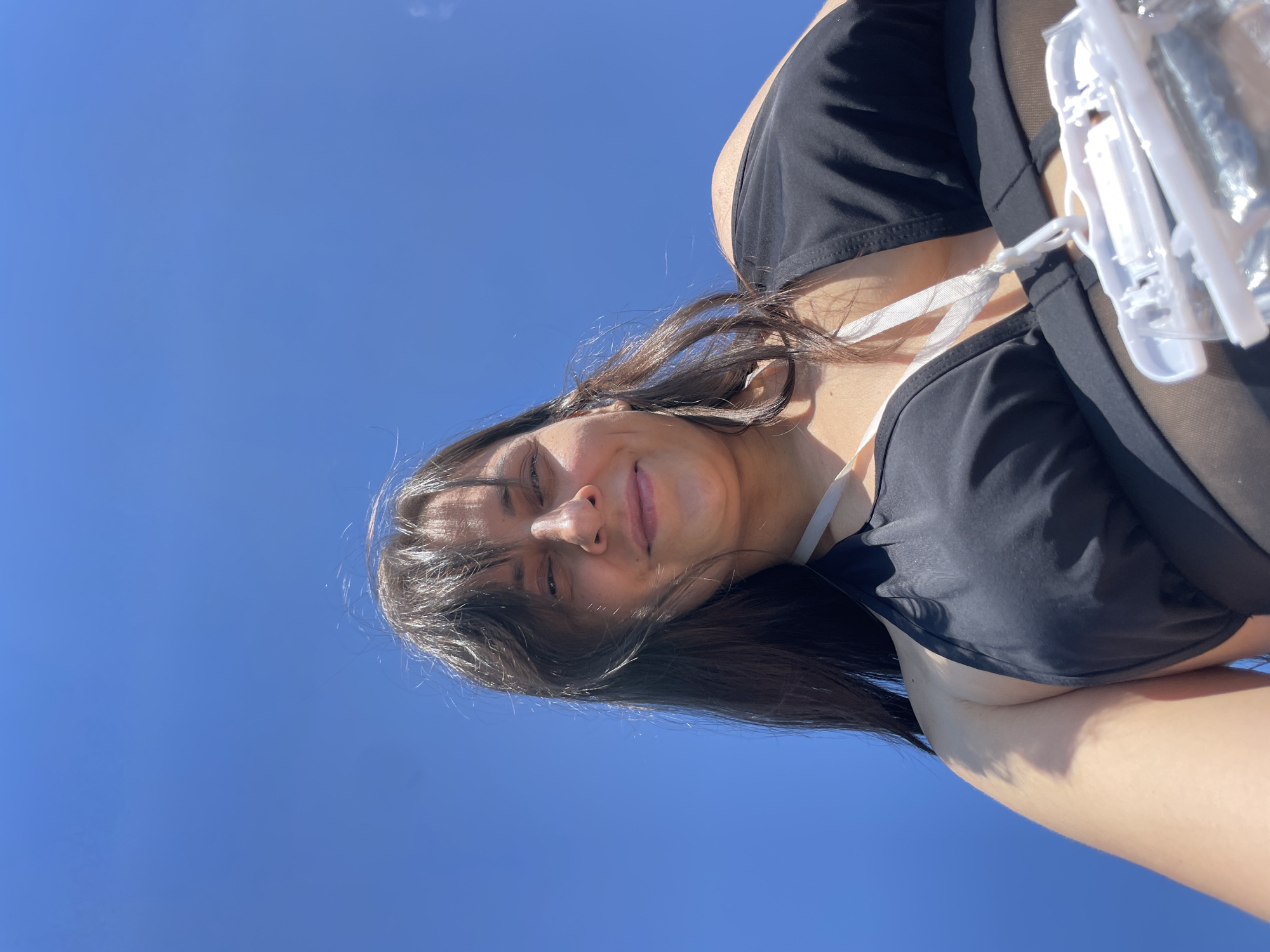 The image is a low-angle shot of a person against a clear blue sky. The person, with dark hair, is wearing a black swimsuit and appears to be looking up at the camera. Part of their face, chest, and shoulder are visible. The background is an unobstructed expanse of blue sky. The image does not contain any information that would allow for the identification of a location in Novyi, Russia.