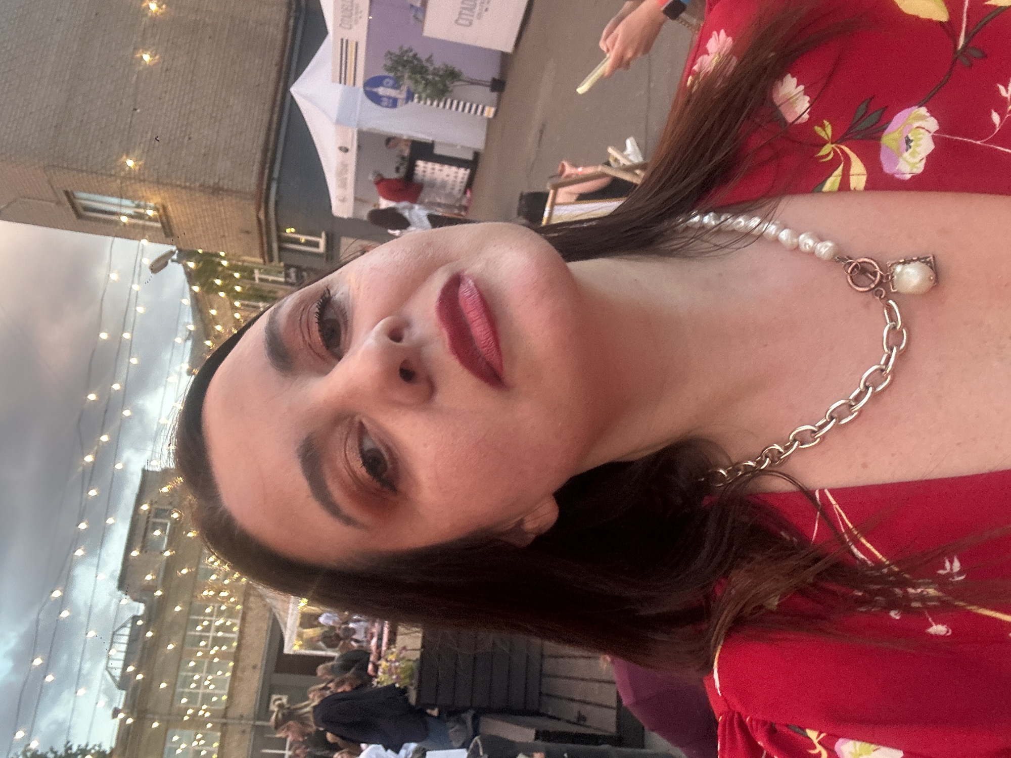 The image depicts a close-up selfie of a woman at an outdoor event in Riga, Latvia, under an overcast sky during late afternoon or early evening.
The primary subject, positioned in the foreground and center, is a woman with dark hair, dark eyes, and fair skin, wearing red lipstick and reddish-brown eyeshadow. She is wearing a red garment with a floral pattern of white, yellow, and pink flowers. A necklace is visible around her neck, featuring a silver-colored chunky chain connected to a string of irregular pearls by a rose gold-colored, circular clasp. A small, square charm containing a single pearl hangs from the clasp. She is looking directly at the camera with her head slightly tilted upward.
The background reveals an urban environment characteristic of an outdoor gathering or festival. Numerous string lights are suspended across the scene and draped on the surrounding buildings. To the left, a multi-story building with numerous windows and an older architectural style is visible. To the right, a brick building also features windows. White event tents are set up in the mid-ground. One prominent tent on the right displays the name "CITADELA" and a logo resembling a stylized building or fortress, confirming the location in Riga, Latvia, given Citadele Bank's headquarters there. A tall, black and white striped structure is positioned near this tent, and a small potted plant stands nearby.
Various people are visible in the background, populating the event space. On the left, figures are partially obscured by a dark, possibly wooden, barrier or planter. Further right, near the tents, individuals are standing, including one person in a red shirt near a counter under a tent. On the far right, a person is seated in a striped deck chair, holding a light-colored smartphone. The ground appears to be a paved or hard surface.