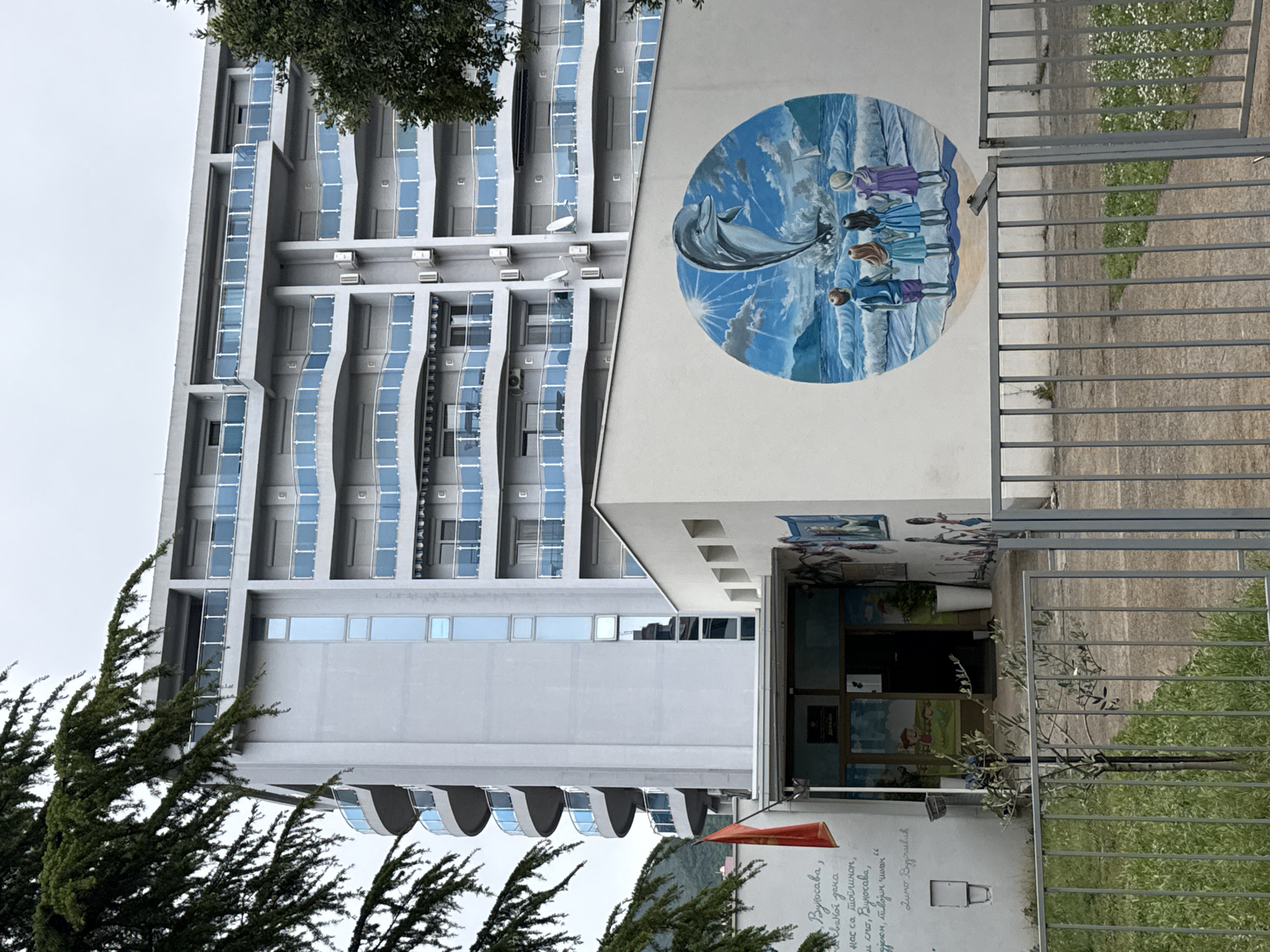The image shows a modern building in Bar, Montenegro, featuring a large, round mural on its side depicting children and a dolphin by the sea. The building, likely a school or community center, has a light gray facade with distinctive vertical window arrangements. A gate stands in front of the entrance, and greenery is visible in the foreground. A red flag with inscription is on the bottom left.