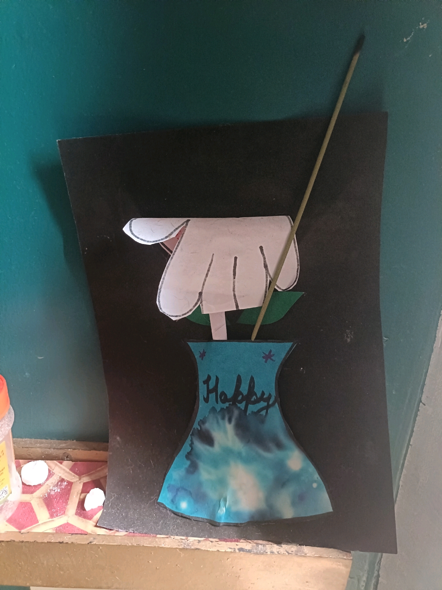 This image depicts a handmade craft featuring a white paper flower in a blue watercolor vase. The word "Happy" is written in cursive on the vase. A thin, light-colored stick, possibly an incense stick or a skewer, is placed behind the flower and extends upwards. The craft is mounted on a black rectangular piece of paper, which is affixed to a teal-colored wall. The lower part of the image shows a wooden shelf with some white powdery material and small white round objects. A partially visible plastic container with an orange lid is also present on the shelf. This scene is likely in Mohanlalganj, India, based on the provided information.