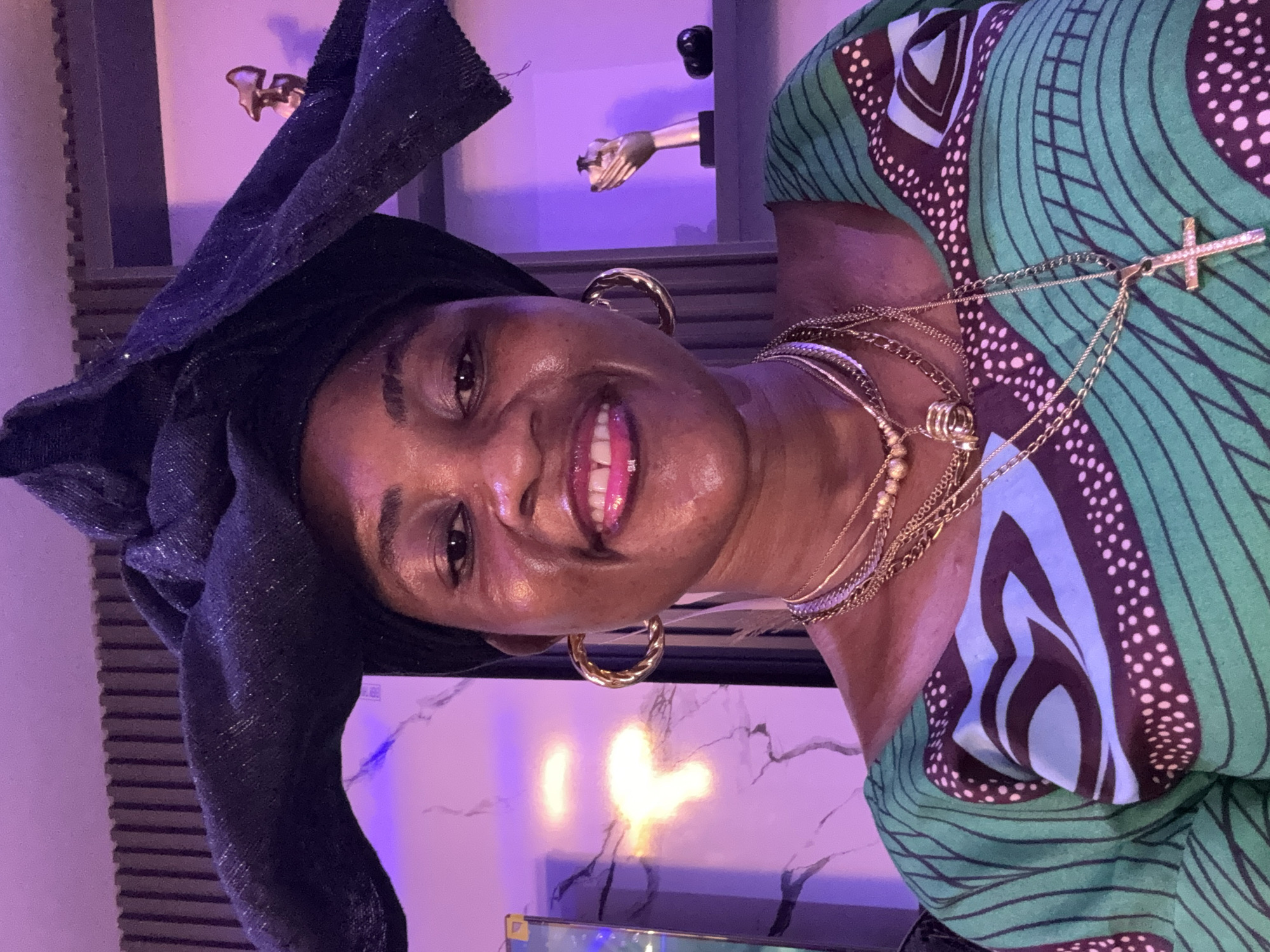The image is a medium shot of a Black woman posing for a selfie. She is wearing a black headwrap with a sparkly texture, large gold hoop earrings, and several gold necklaces, including one with a diamond-encrusted cross pendant. She is also wearing a green dress with a geometric pattern in blue, brown, and white. The woman is smiling, with her teeth showing, and has on pink lipstick. She is indoors, in what appears to be a room with purple lighting. Behind her, shelves are visible with decorative objects, including a golden hand sculpture. The room's decor includes a textured wall and a marbled pattern. The image is focused on the woman's face and upper body. There is no information in the image to suggest the location is in the city of Ajara, Nigeria.