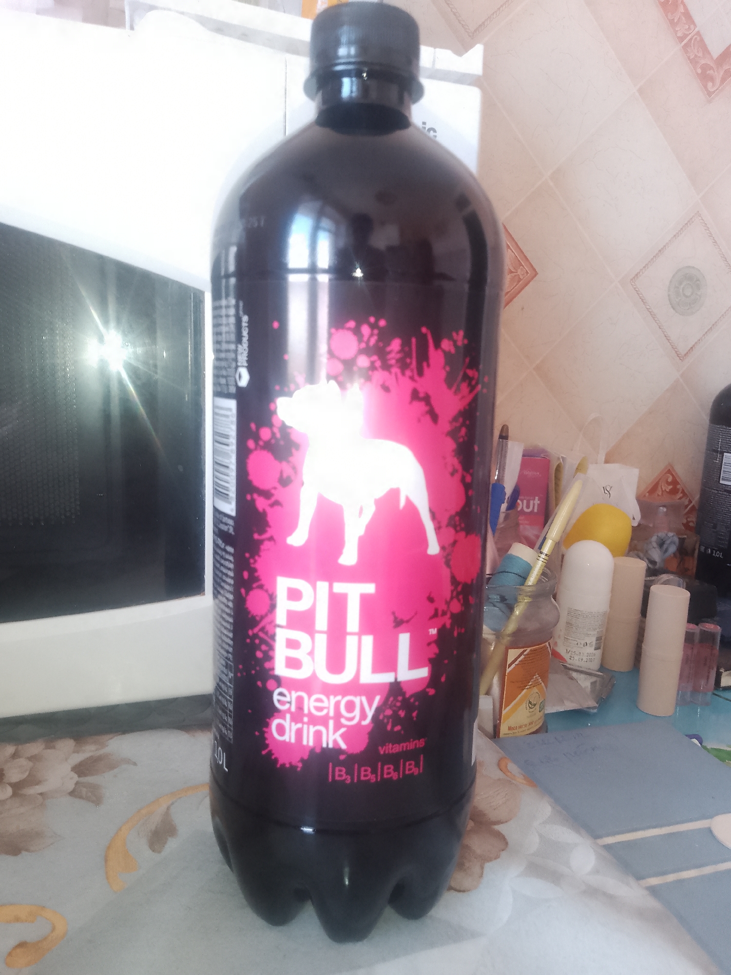 A dark plastic bottle of Pitbull energy drink with a pink label featuring a white pitbull silhouette and pink splatter is the main subject of the image. The bottle is positioned in the foreground, slightly to the right of center, on a surface covered with a patterned tablecloth. In the background, to the left, is a portion of a white appliance, possibly a microwave, with a reflective surface. To the right, the background consists of a tiled wall and a cluttered tabletop with various items including pens, a yellow object (possibly a lemon), and several cylindrical containers. The lighting suggests an indoor setting. The location is identified as Chernivtsi, Ukraine.
