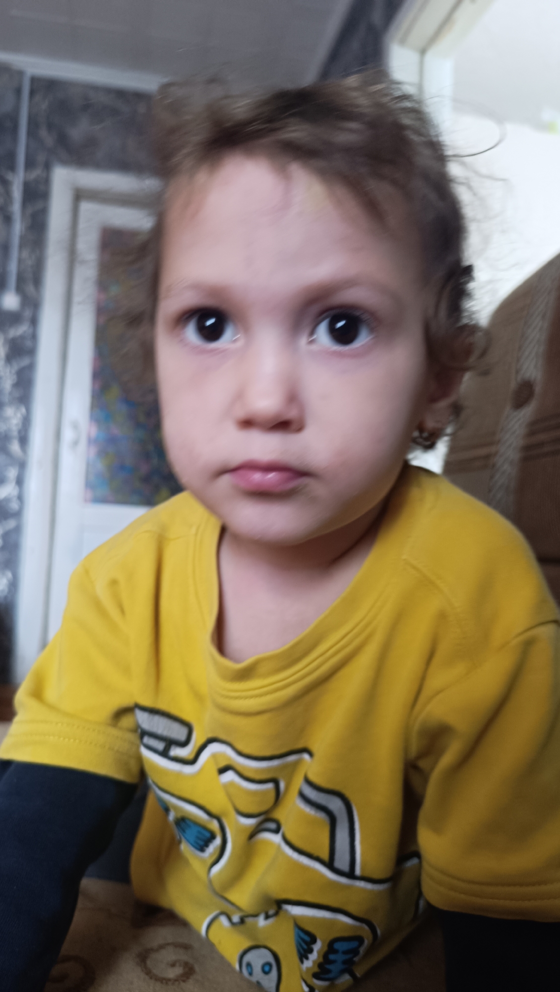A young child with light brown, curly hair and dark eyes is pictured from the chest up. The child is wearing a bright yellow t-shirt with a graphic design in white, grey, and blue. The child's left arm is covered by a dark blue sleeve. The background is out of focus but appears to be an indoor setting with textured wallpaper and a doorway. The image is a close-up portrait of the child, who is looking directly at the camera with a neutral expression. The lighting suggests an indoor environment, possibly a home. There are no discernable elements within the image that indicate a specific location in the city of Karsha, Russia.