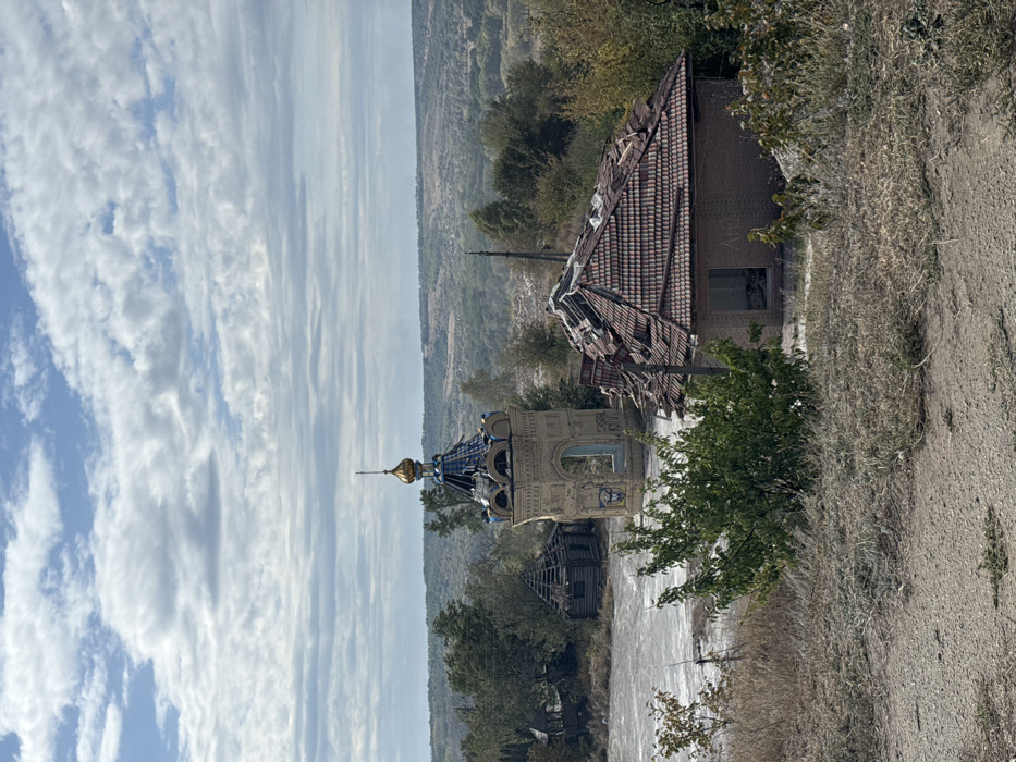 This image depicts a damaged landscape in Izium, Ukraine, specifically around the Holy Spring of the Pishchanska Icon of the Mother of God. The scene is observed from an elevated, unpaved vantage point, characterized by dry, sparse vegetation in the immediate foreground.
In the midground, a light-colored, octagonal brick or stone chapel structure with a blue and gold onion dome stands prominently. The dome exhibits significant damage, with numerous blue tiles missing and exposed structural elements. An Orthodox cross is visible atop the dome. The chapel's arched openings are empty, lacking windows or doors, and some graffiti-like markings are present on its lower walls, including partially visible Cyrillic text and a blue emblem. To the right of the chapel, a single-story brick building with a gabled, red tiled roof is severely compromised; multiple tiles are missing, exposing the underlying wooden rafters, and sections of the roof appear collapsed. An open doorway or window on its façade reveals interior debris. Further to the left and slightly behind the chapel, a smaller, dilapidated wooden hut with a partially collapsed roof is visible. The ground immediately surrounding these structures is light-colored, appearing to be bare earth, rubble, or dried mud. Interspersed among the buildings and in the foreground are various bushes and trees, some green and others exhibiting autumnal brown foliage.
The background consists of extensive rolling hills covered in dense green and brown woodland, stretching to the horizon under a sky with scattered cumulus clouds and patches of clear blue. No people are visible in the image.