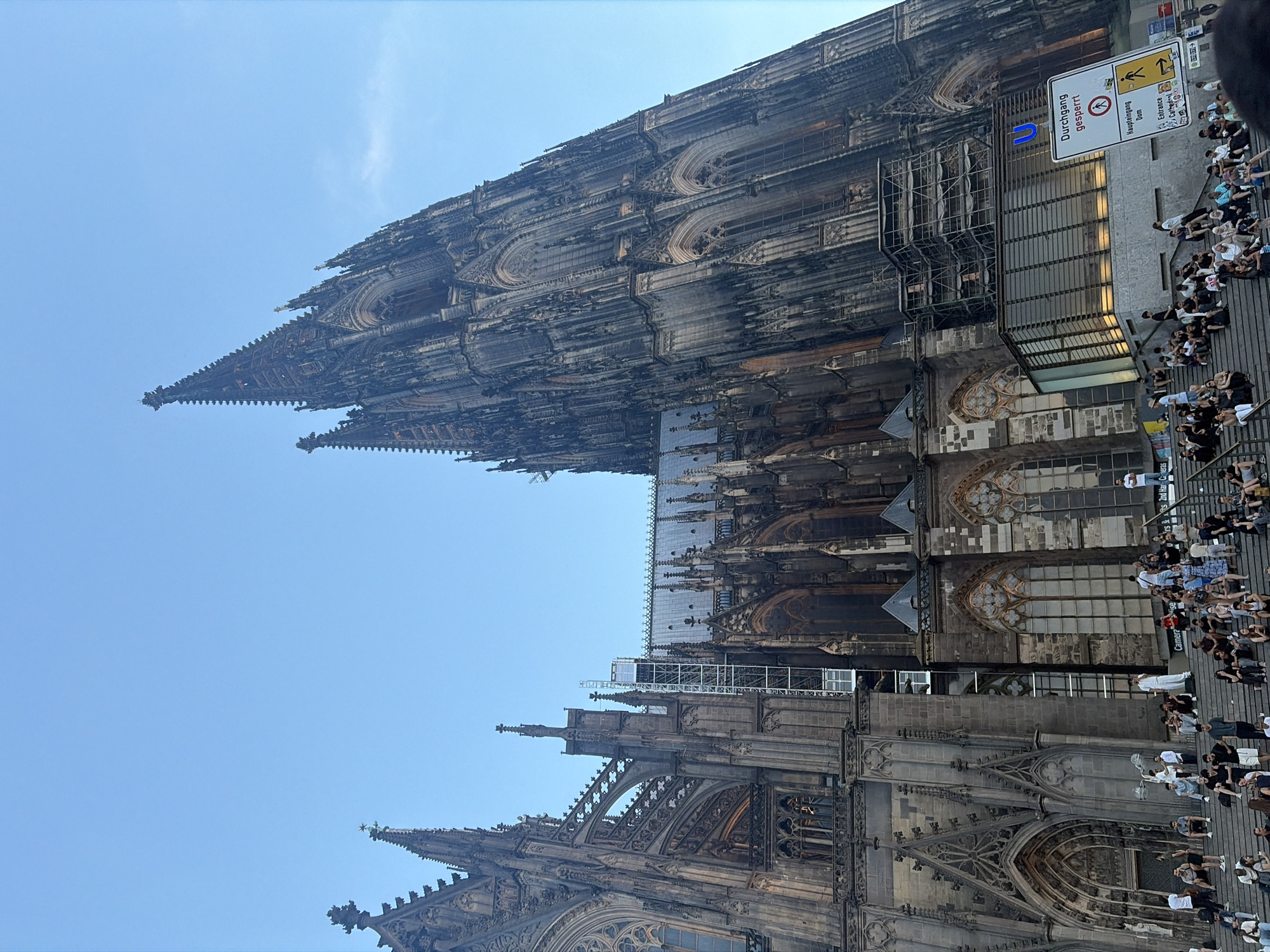 This is a low-angle, vertical, eye-level shot of the Cologne Cathedral in Cologne, Germany, under a clear blue sky. The imposing gothic architecture of the cathedral dominates the frame with its intricate spires, ornate stonework, and stained-glass windows. Scaffolding is visible on the left side of the building, suggesting ongoing restoration or maintenance. In the foreground, a wide set of stone stairs is filled with a diverse group of people sitting, standing, and interacting. They appear to be tourists and locals enjoying the late afternoon or early evening. Some are taking photos, while others are engaged in conversation or simply relaxing. A modern glass-enclosed structure with illuminated interior is situated at the base of the cathedral, near the stairs. A signpost with German text and directional arrows is also visible, indicating an entrance or information point. The overall atmosphere is one of a bustling urban landmark attracting visitors and residents alike.