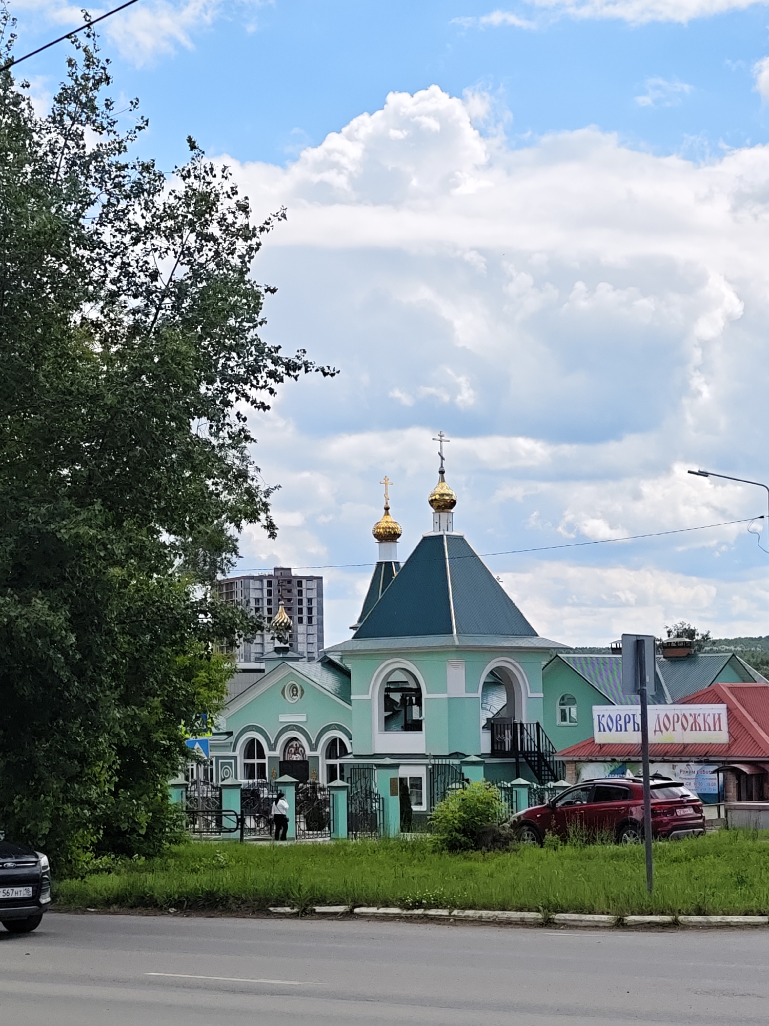 The image depicts a street view in Chaikovsky, Russia, on a partly cloudy day. In the foreground, a paved road with a white line is visible. To the right of the road, a strip of green grass with some small white flowers runs along a curb. Beyond the grass, a red SUV is parked. Further back, a light green building with arched windows and golden domes, typical of Orthodox churches, is prominent. A sign with Cyrillic text, reading "КОВРЫ ДОРОЖКИ," is attached to a building to the right of the church. To the left of the church, a large tree with lush green foliage partially obscures the view. Behind the church, a multi-story building under construction is visible. The sky is blue with scattered white clouds.